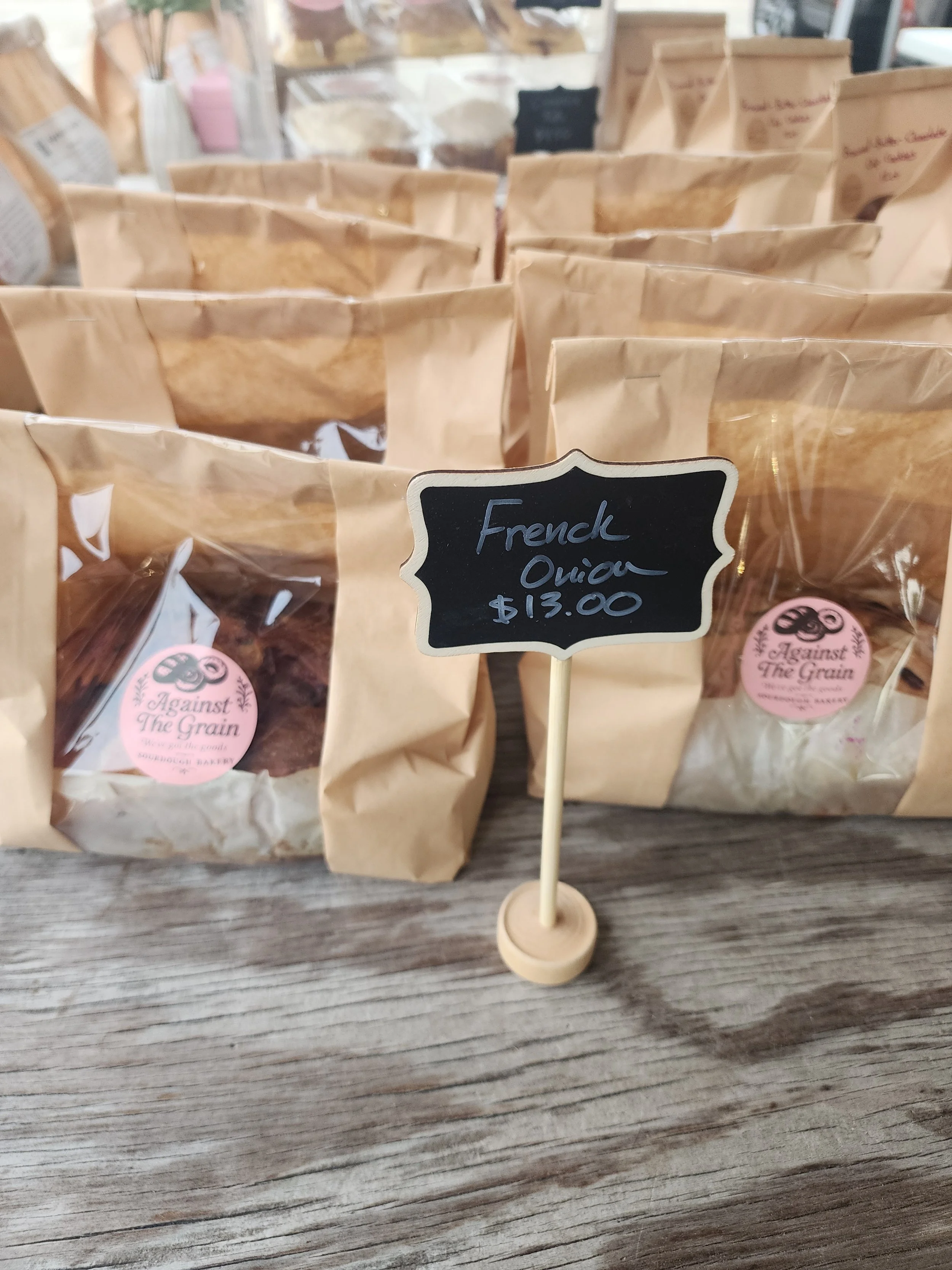 Close-up of a display of brown paper bags labeled 'Against The Grain' with a chalkboard sign reading 'French Onion $13.00' in front of them, on a rustic wooden table.