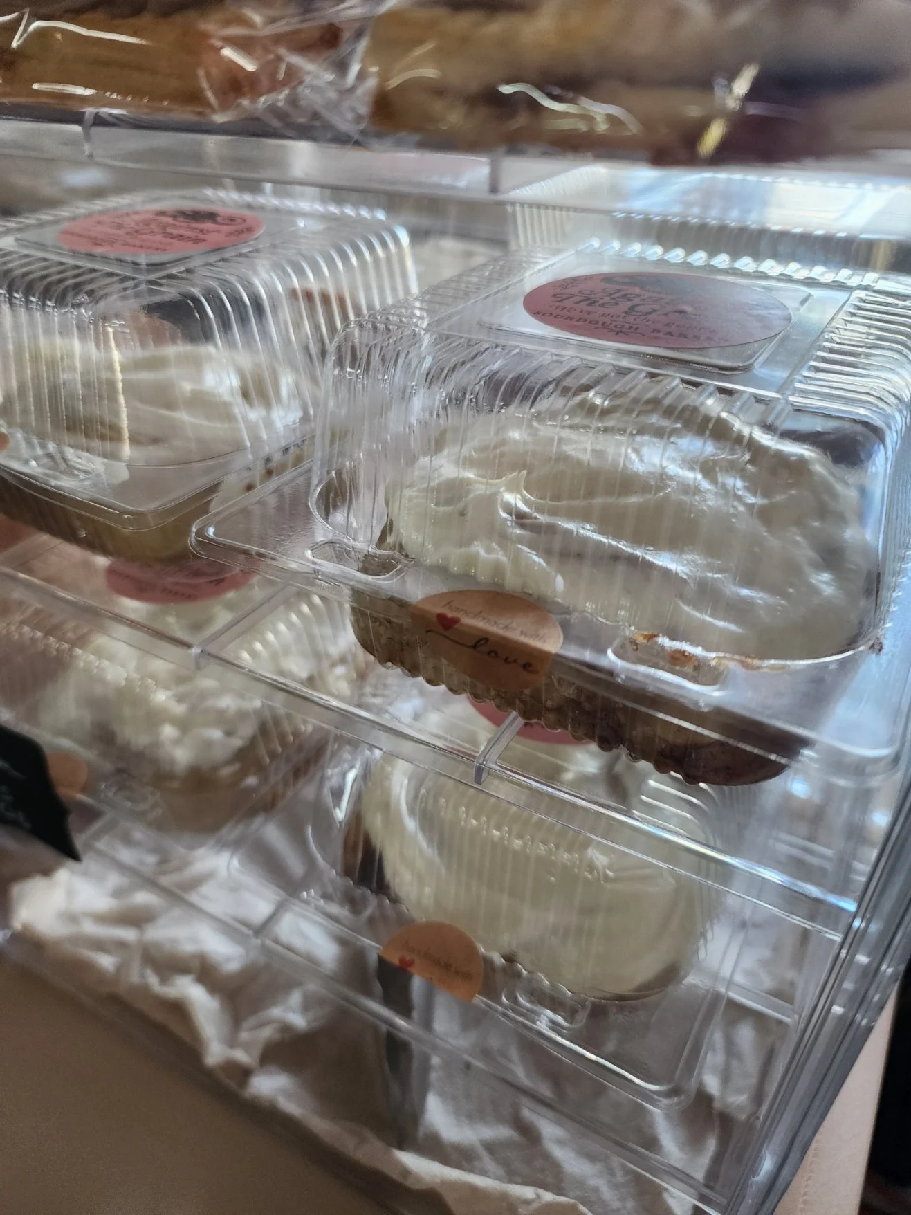 Inside a refrigerator, there are multiple clear plastic containers of assorted desserts, including cakes or cheesecakes, with pink and beige heart-shaped labels.