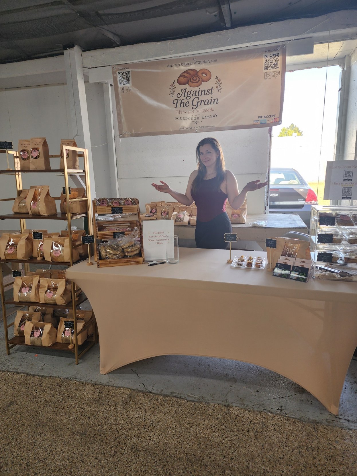 A woman at a bakery booth with a banner reading "Against The Grain" and displays of baked goods and bags of bread.