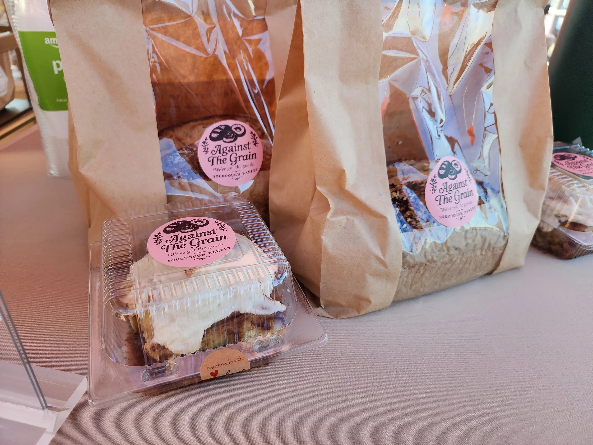 Brown paper bags with a circular pink label that reads 'Against The Grain, We've got the goods, SOURDOUGH BAKERY.' Clear plastic containers with sliced cake. Part of a green and white package visible on the left. The items are on a light-colored tabl