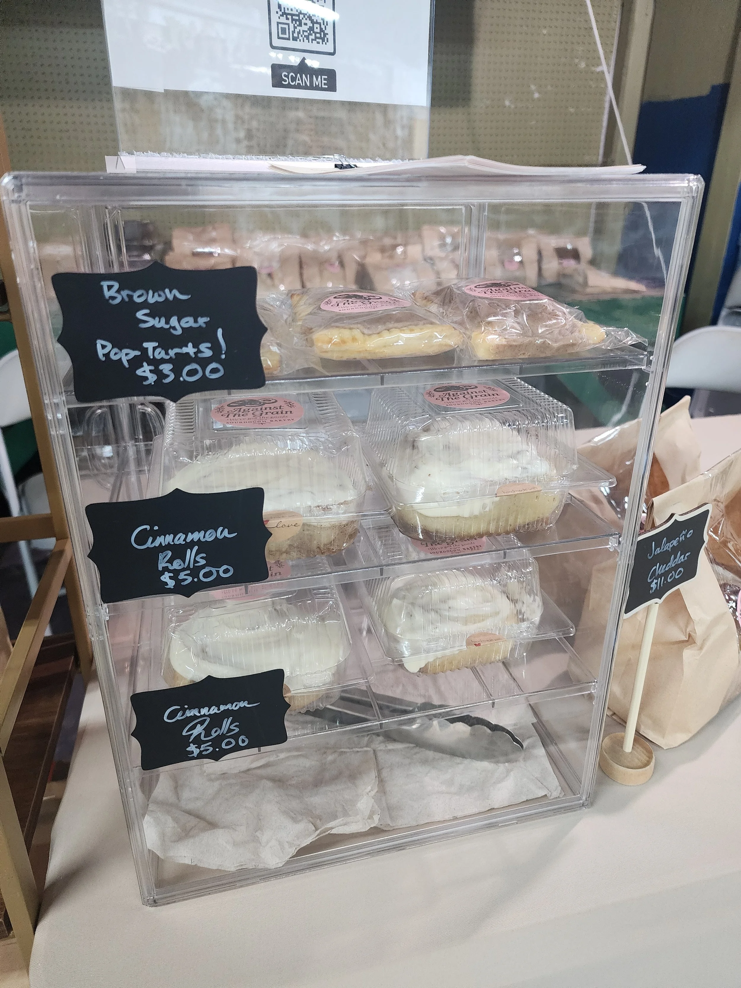 A clear acrylic display case containing various baked goods, including cinnamon rolls and other pastries, with handwritten chalkboard signs indicating prices for brown sugar pop-tarts and cinnamon rolls.
