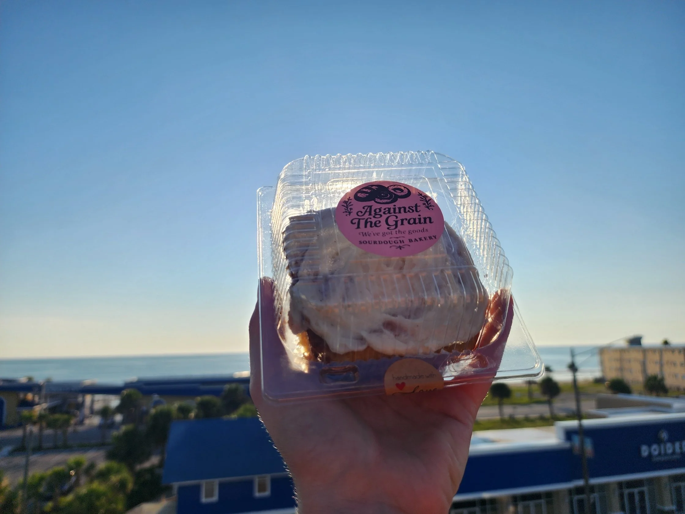 A clear plastic container of sourdough baked goods held up against a blue sky, with a cityscape and ocean in the background.