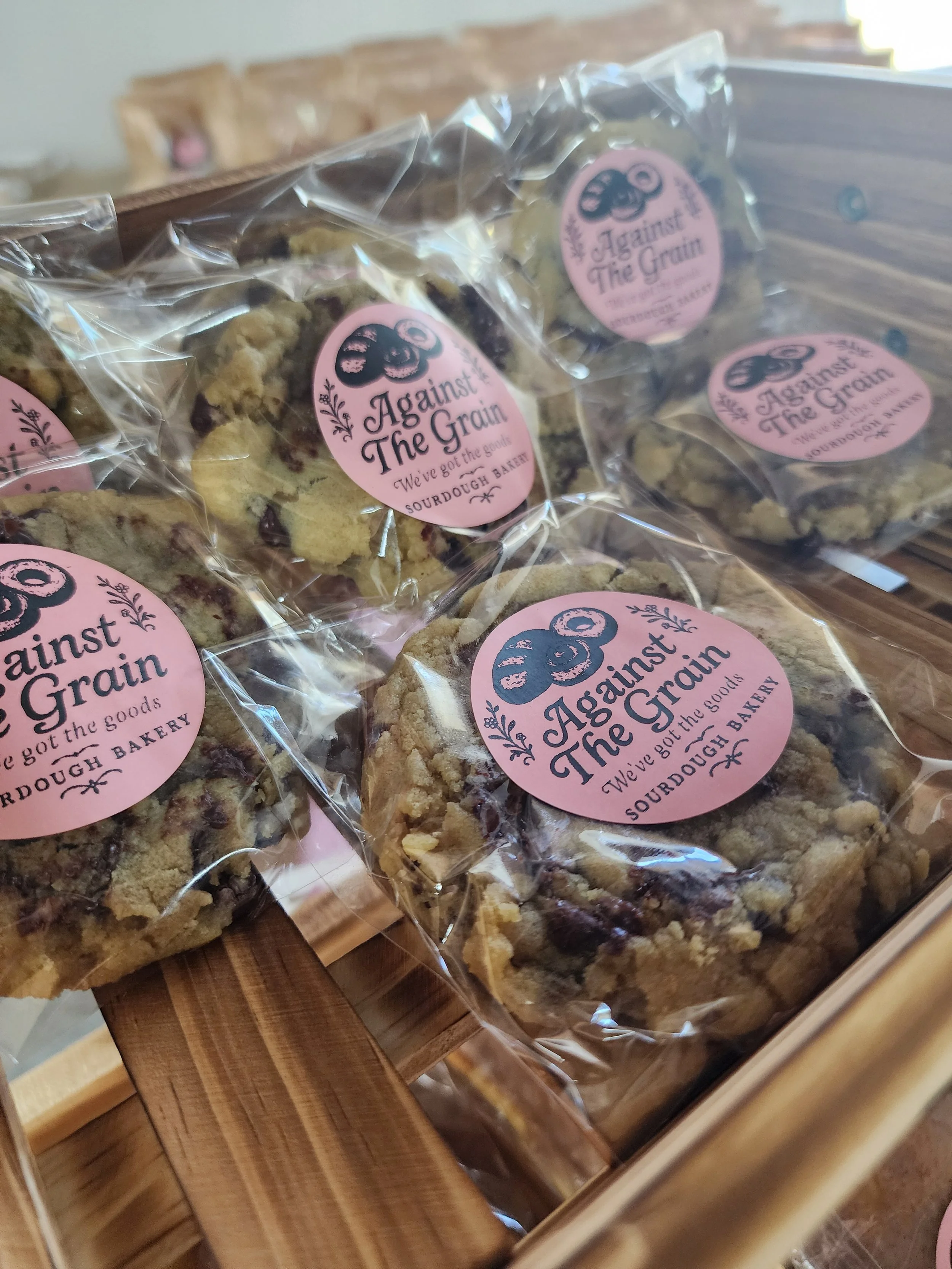 Clear plastic bags of cookies with pink labels reading "Against the Grain," placed on a wooden surface.