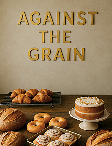 A table with various baked goods including croissants, bagels, cinnamon rolls, and a cake with white frosting. In the background, on the wall, there are gold 3D letters spelling out "AGAINST THE GRAIN".