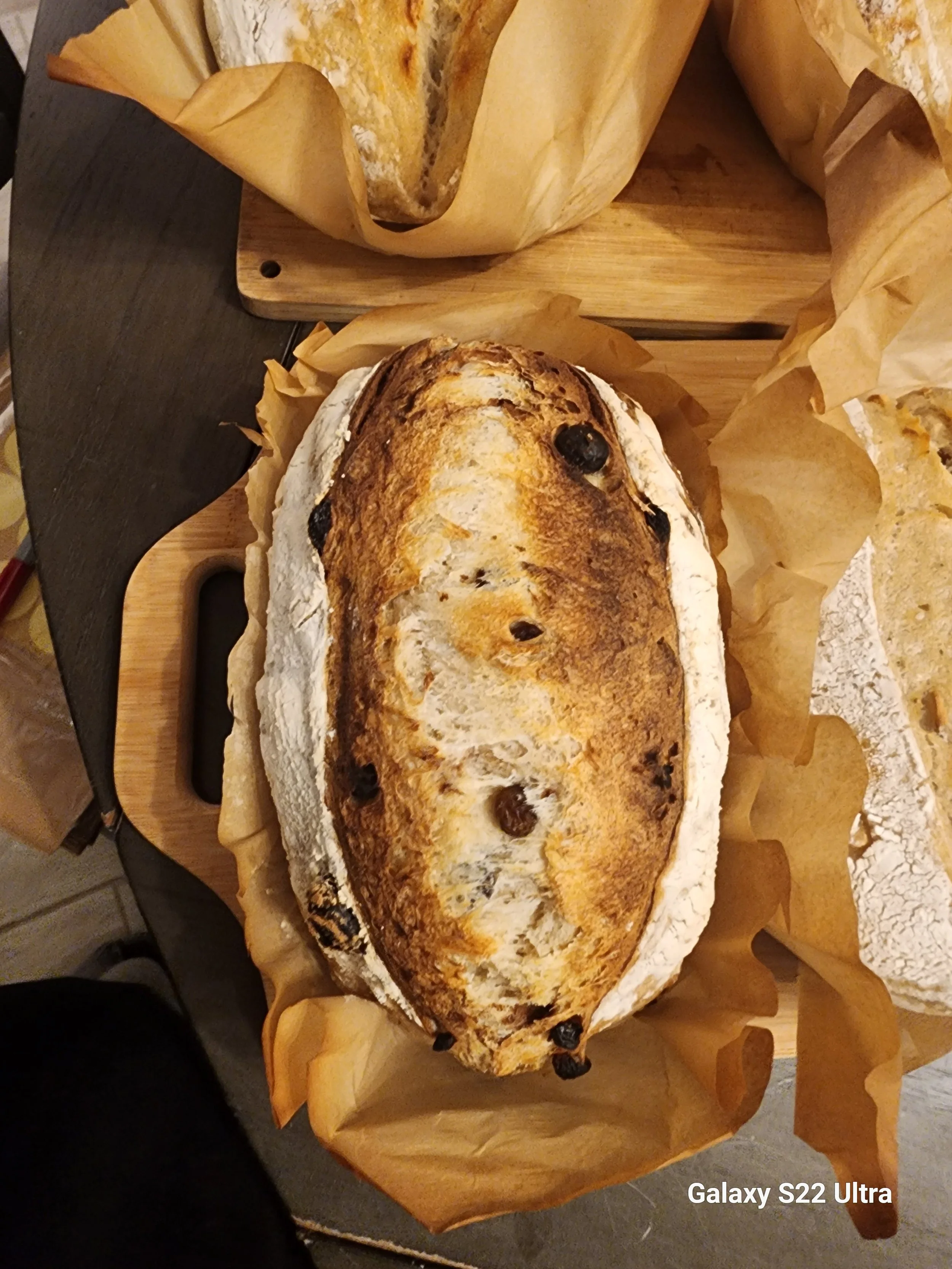 A loaf of bread with a golden-brown crust and a slightly burnt appearance, resting on parchment paper on a wooden cutting board, with a black dried fruit or berry at one end.