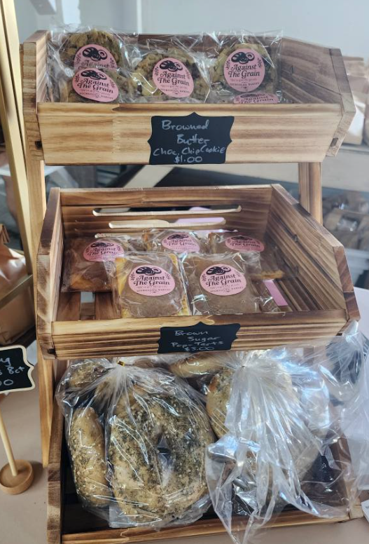 Wooden display stand with packaged baked goods, including cookies and bagels, labeled with pink and black stickers and chalkboard tags.