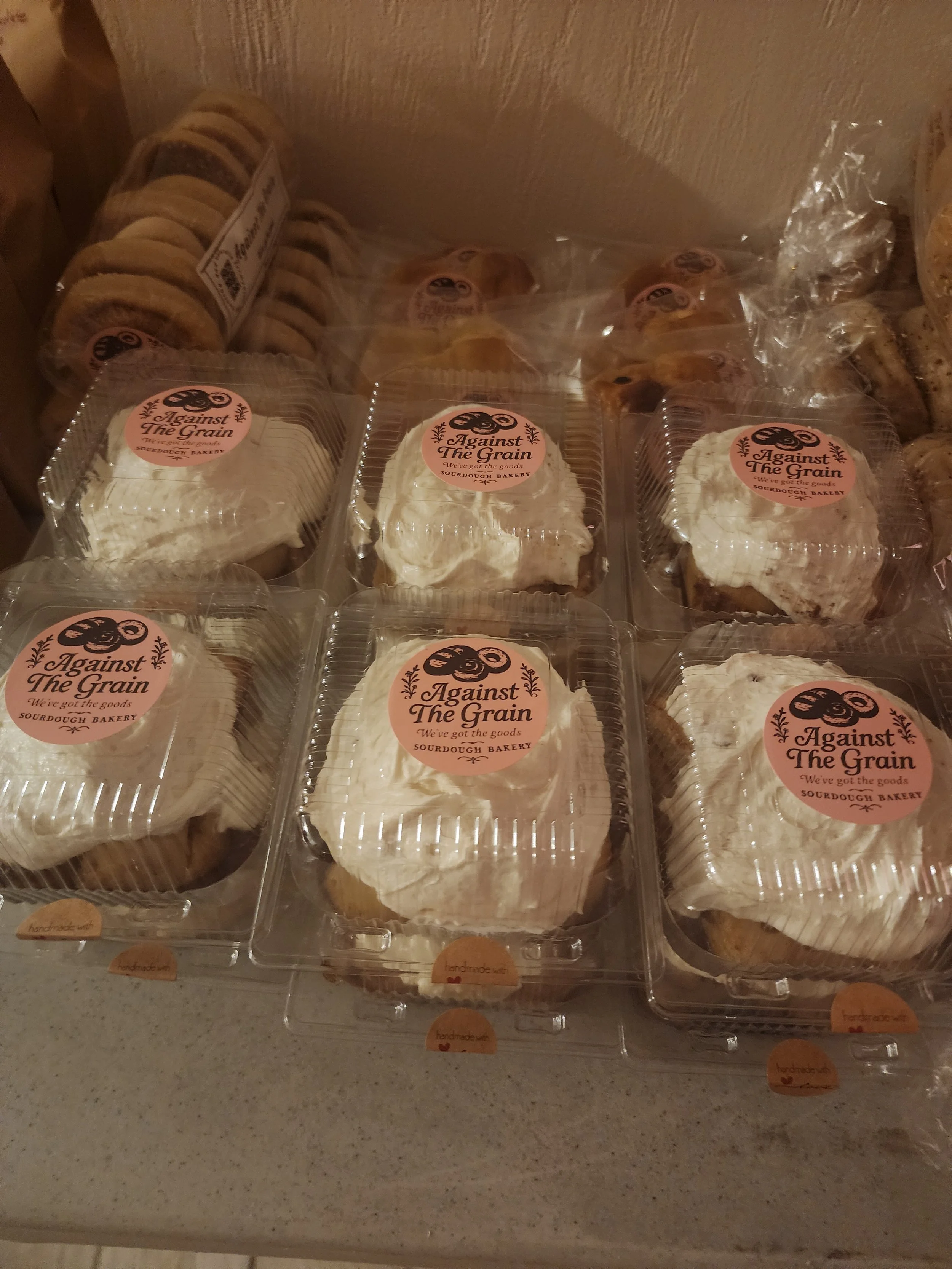 Plastic containers of baked goods labeled with "Against The Grain" and "We're got the goods," featuring cupcakes topped with white frosting and sugar cookies on a countertop.