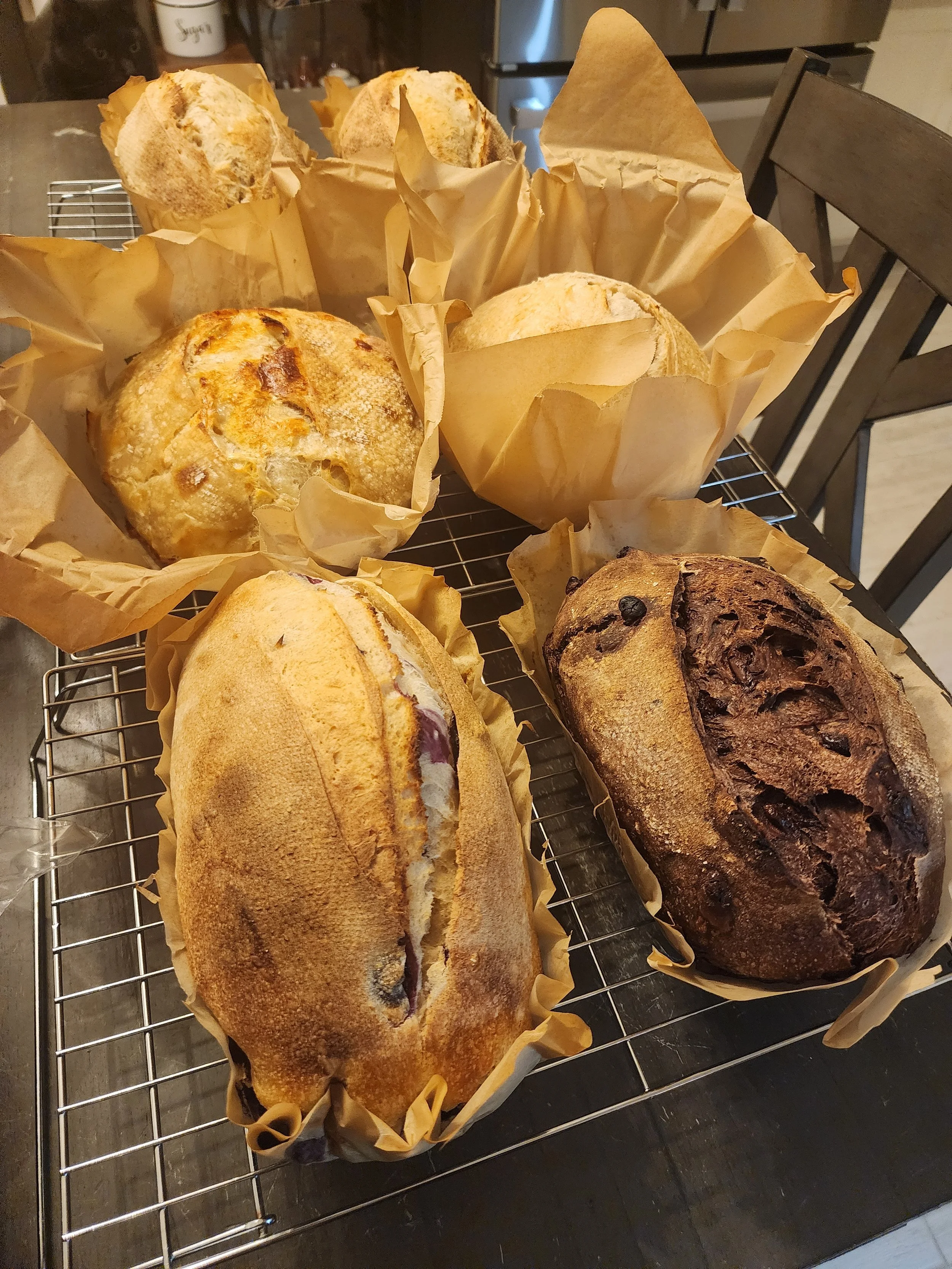 Various loaves of bread cooling on a wire rack, including a rustic round loaf, a sliced baguette, and a darker, crusty loaf, all wrapped in parchment paper.