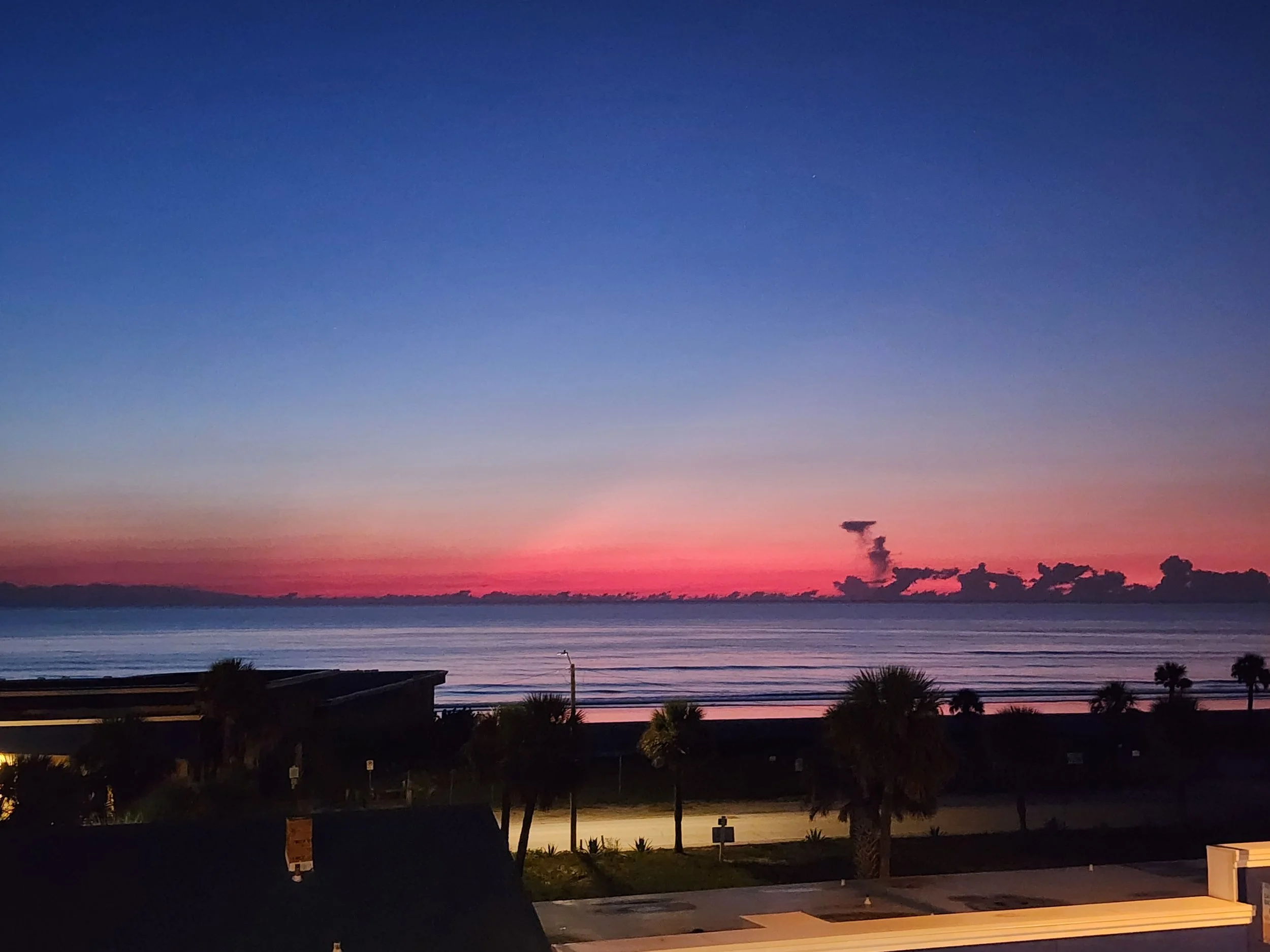 Sunset over the ocean with a colorful sky in shades of pink, purple, and blue, and silhouettes of trees and buildings in the foreground.