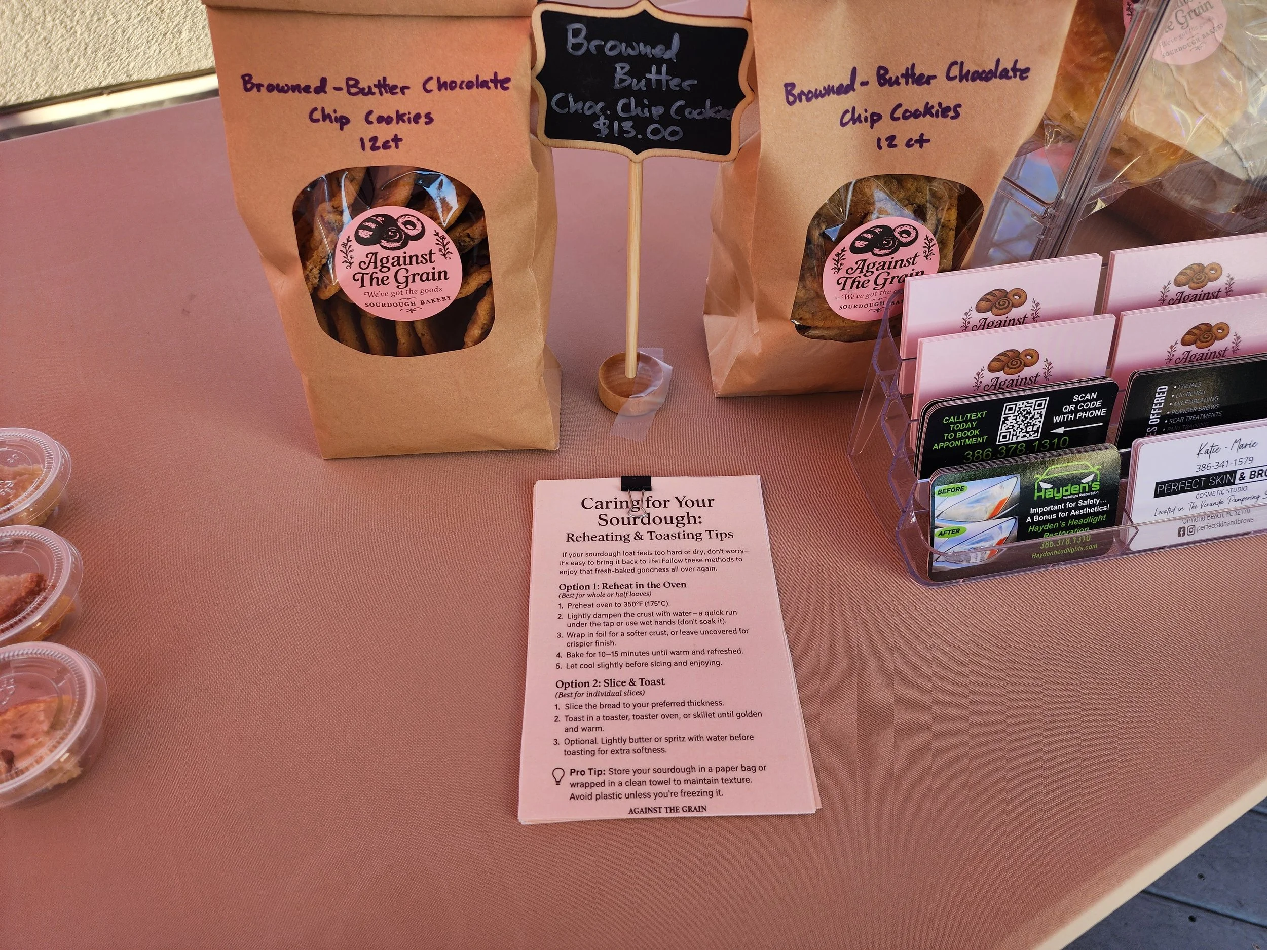 Bags of browned butter chocolate chip cookies on display with handwritten signs and informational paper about reheating and toasting sourdough bread.