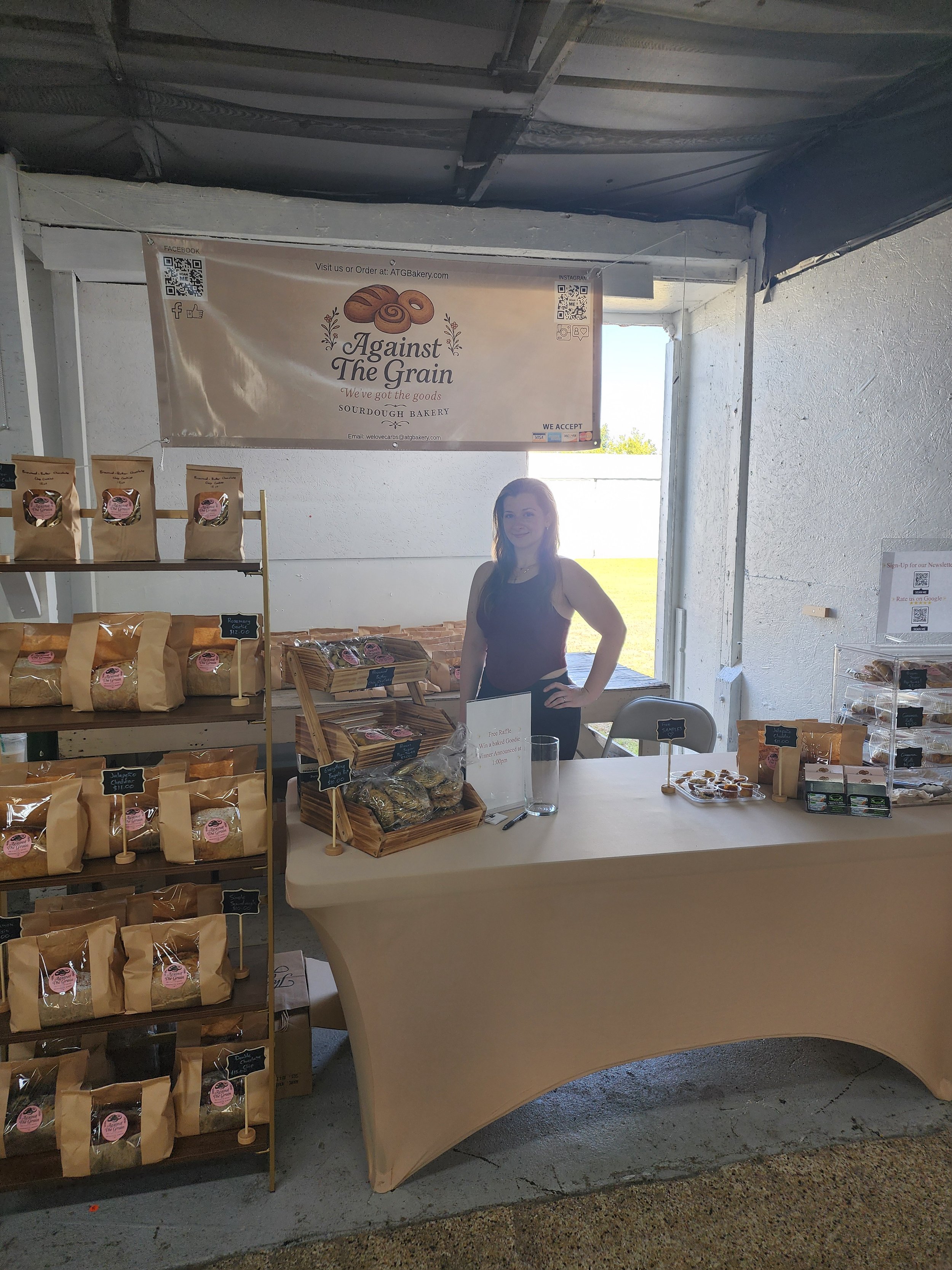 A woman standing at a bakery booth inside a white tent, with a banner that reads "Against The Grain" and displays baked goods, organized on shelves and a table.