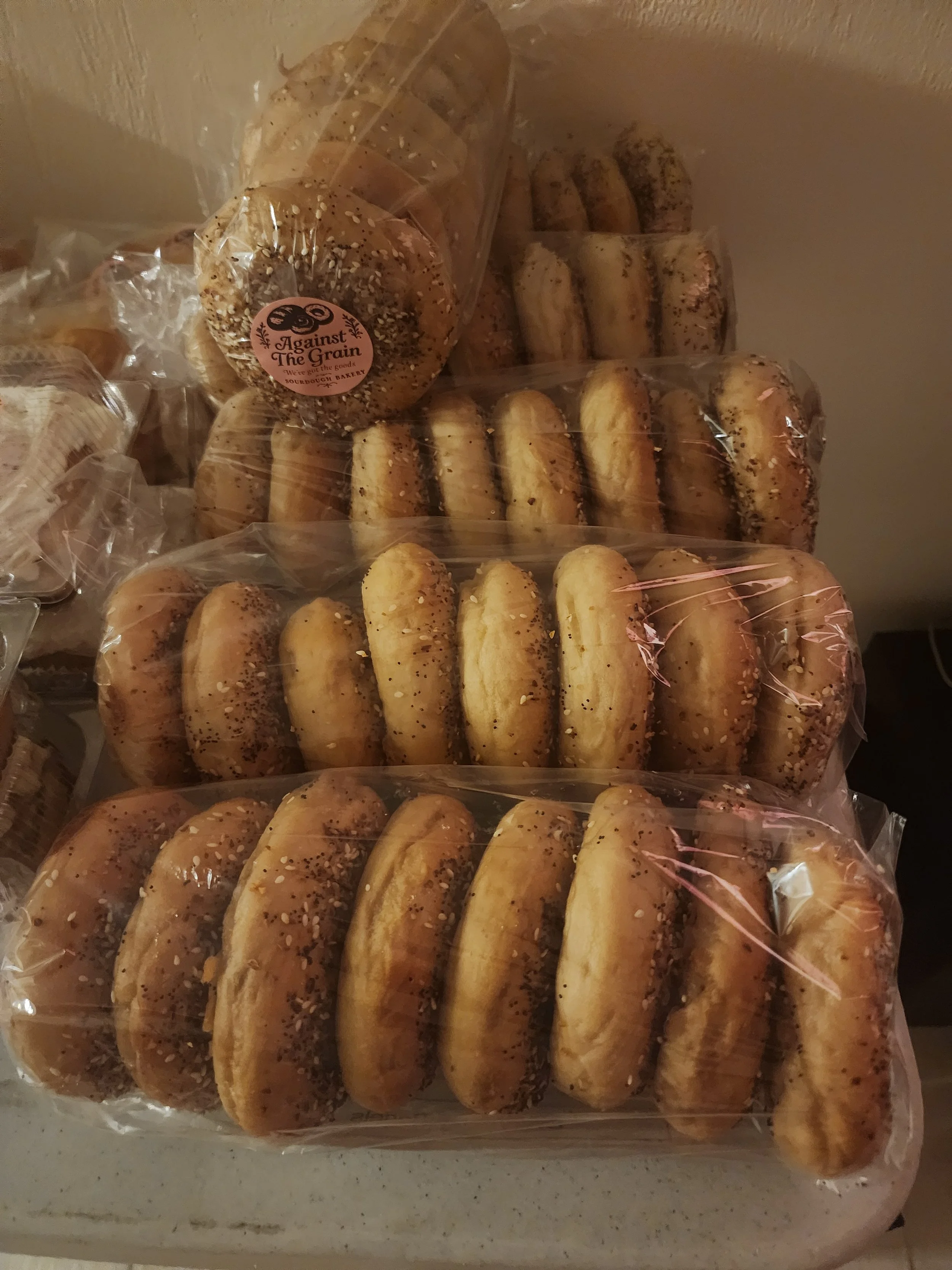 Stacked bagels in plastic packaging on a countertop, with an 'Against The Grain' sourdough bakery sticker visible on one bagel.