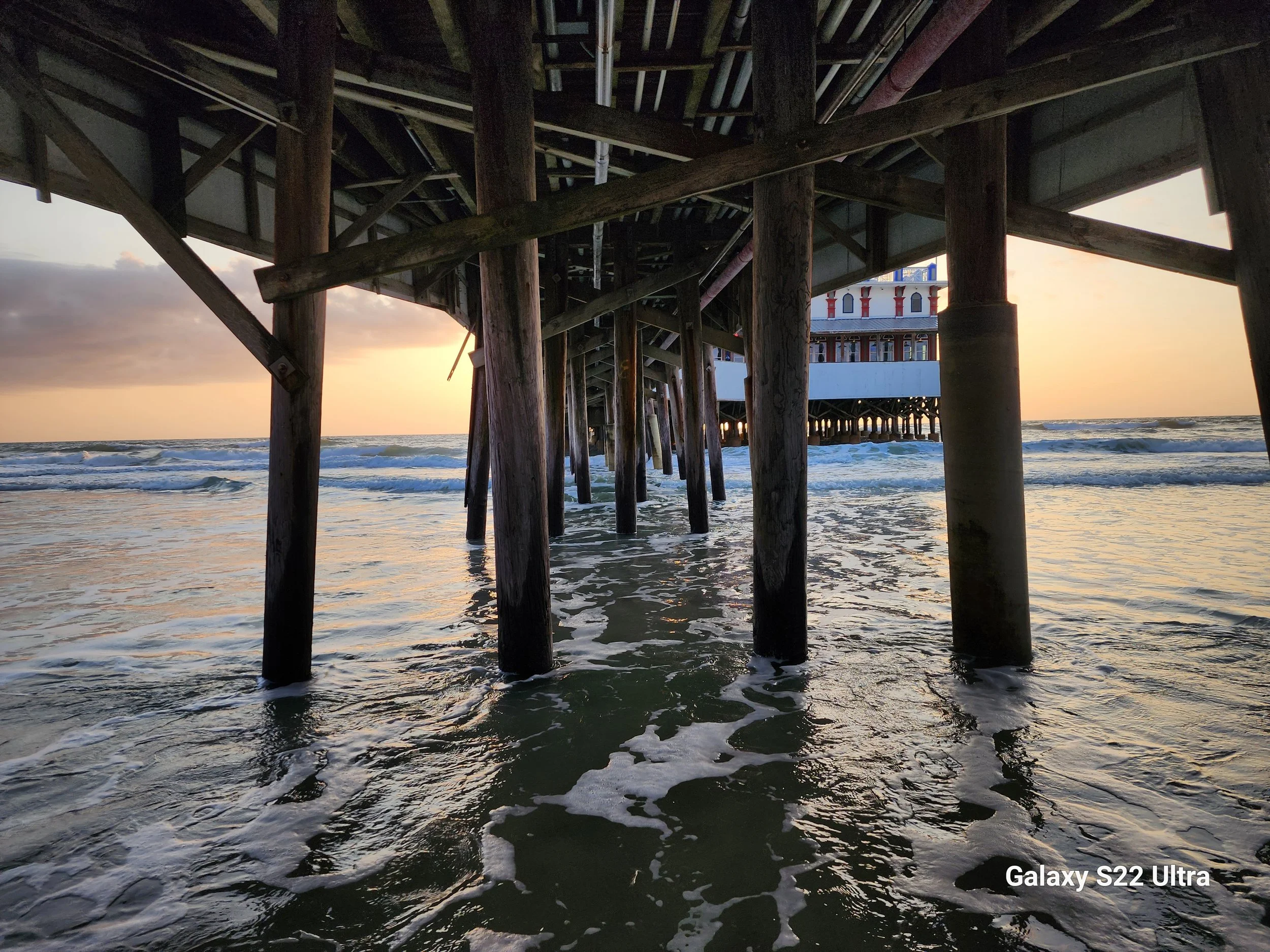 Underneath a pier with wooden supports at sunset, the ocean waves are visible with a building on the pier in the background.