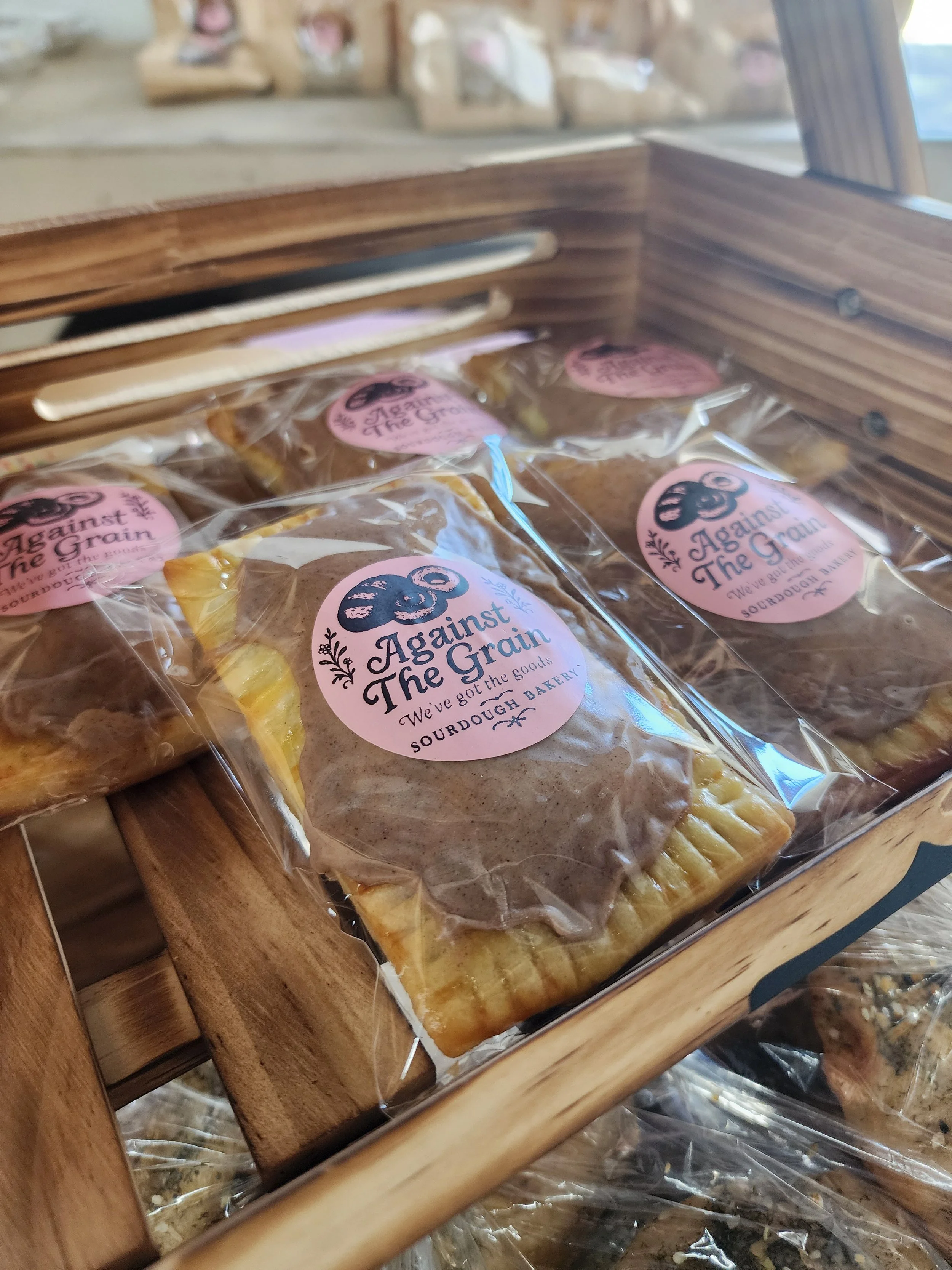 Plastic-wrapped baked goods on a wooden display shelf at a bakery, with a pink circular sticker that reads "Against The Grain" and "We’ve got the goods".