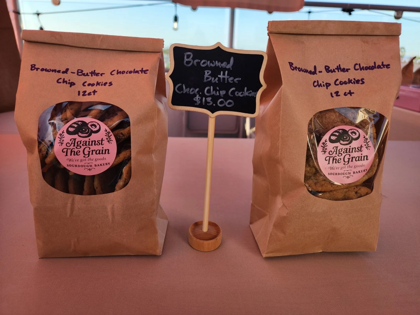 Two brown paper bags of chocolate chip cookies from Against the Grain Bakery, with a black chalkboard sign in the middle reading "Browned Butter" and a price of $13.00.