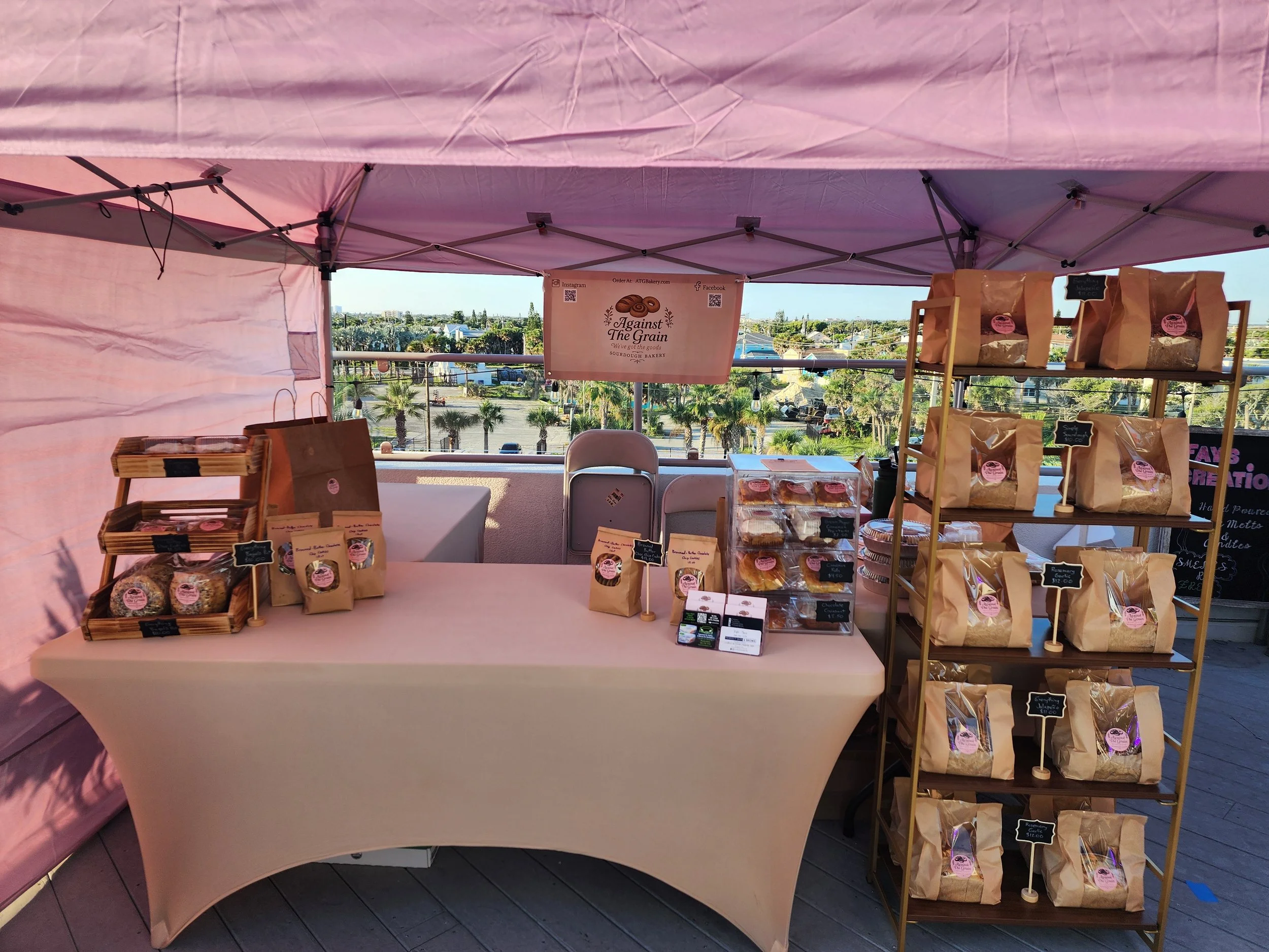 A market stall with baked goods, featuring a pink canopy and shelves filled with brown paper bags of cookies and treats, with a cityscape and palm trees in the background.