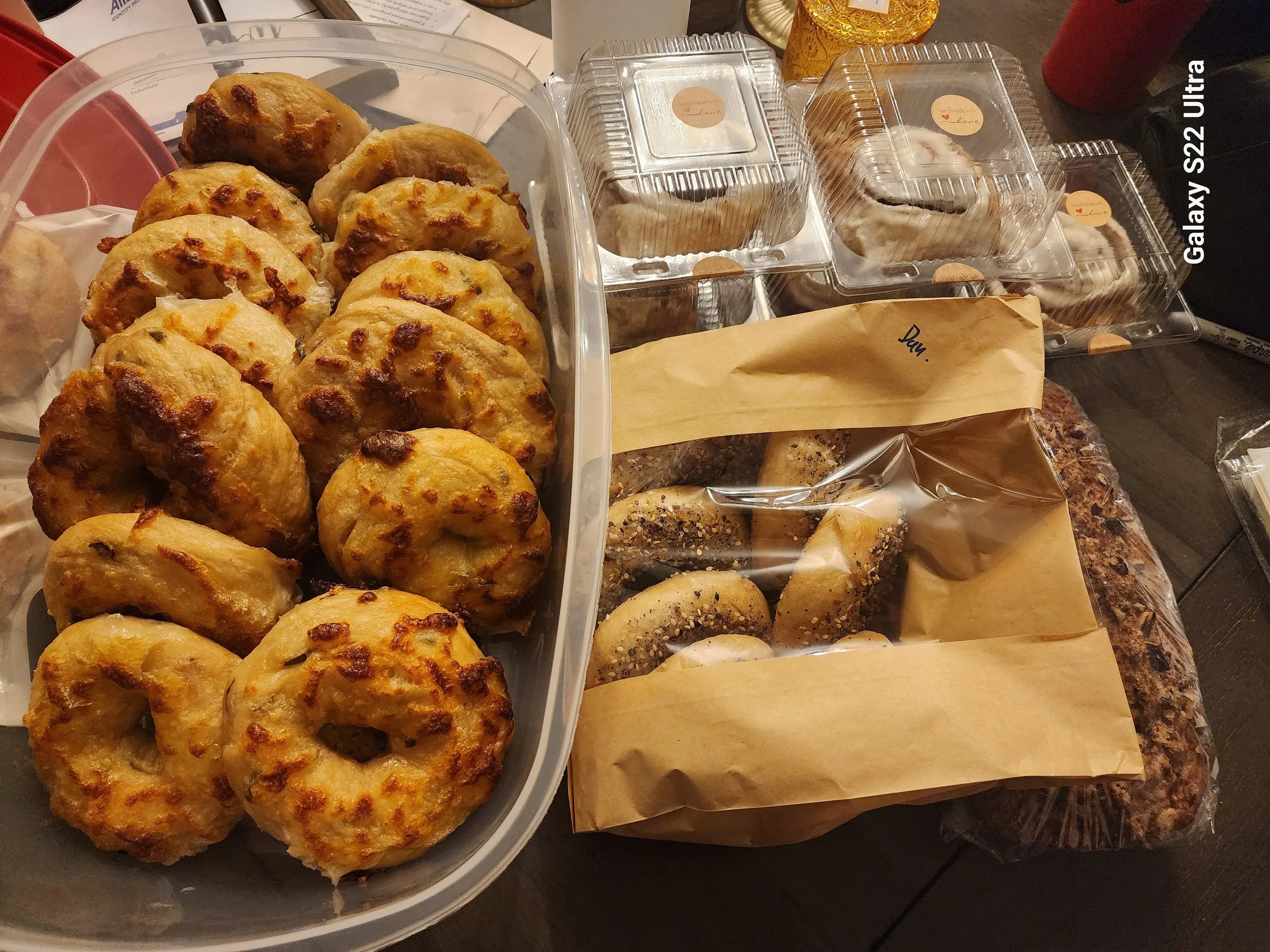 A plastic container filled with baked bread rolls with cheese and herbs. Next to it are several clear plastic containers with baked goods, each labeled with stickers that say "handmade with love." To the right, there are more baked items, some with s
