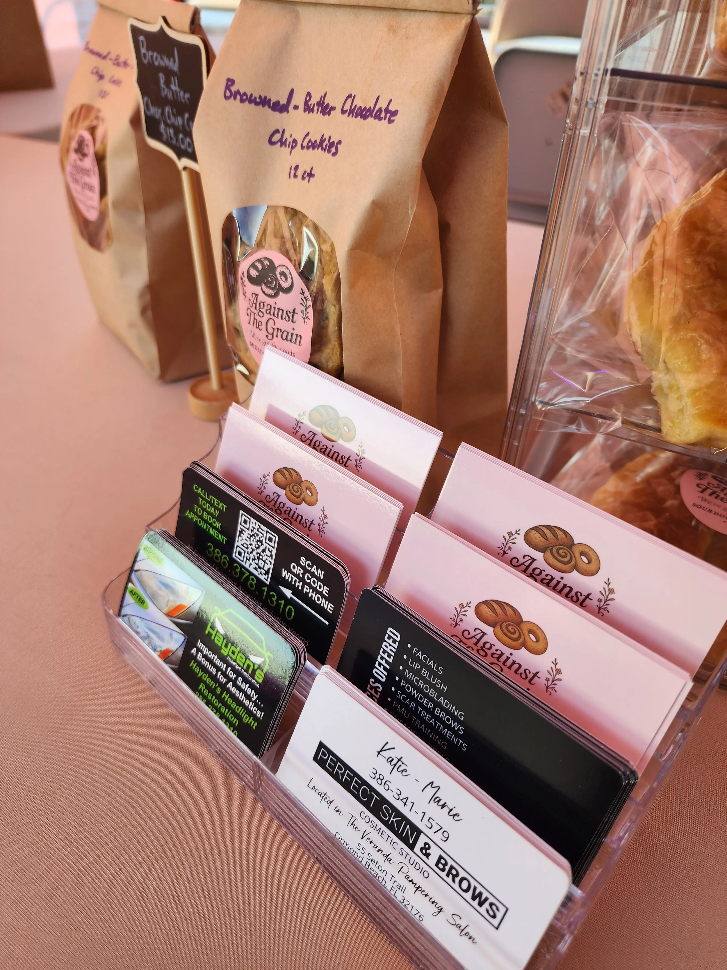 Clear plastic container with business cards and brochures promoting a skincare and brow salon, with a paper bag of cookies labeled "Ground - Butter Chocolate Chip Cookies 12 ct" in the background.