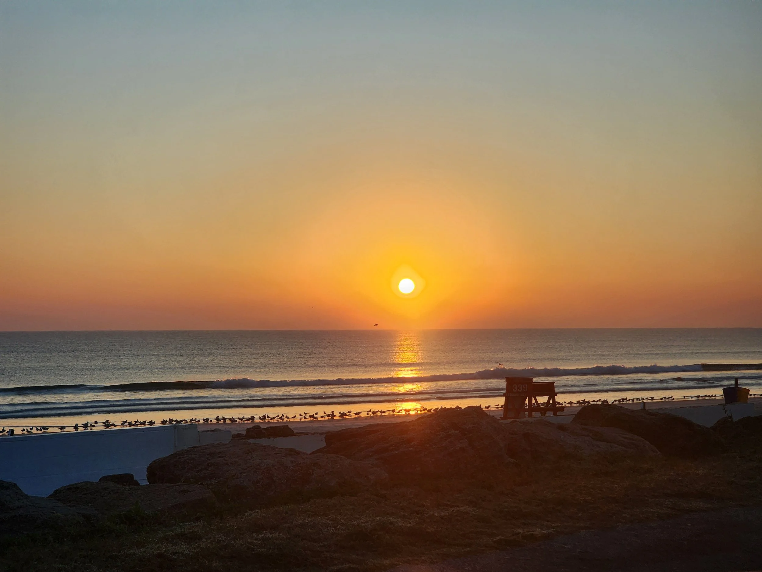 A sunset over the ocean with colorful sky, birds on the beach, rocks, and some structures in the foreground.