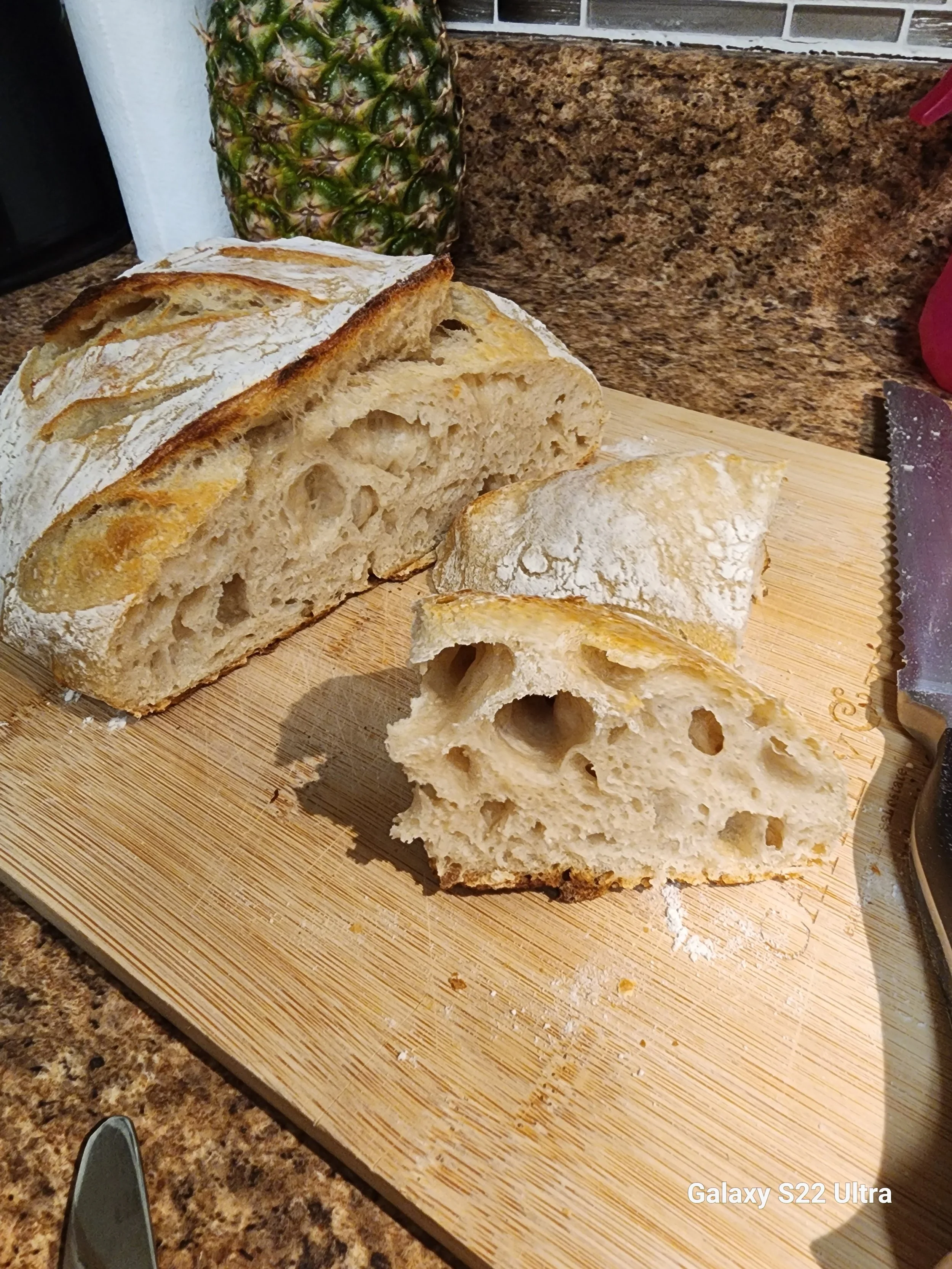 A loaf of bread on a wooden cutting board with some slices cut and separated, a pineapple in the background, and a serrated knife to the right. The countertop is made of a speckled brown material.