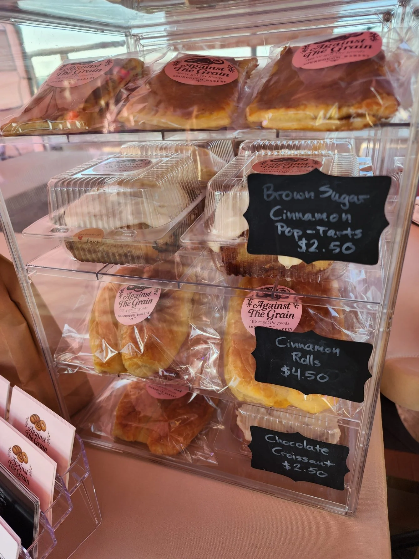 A display case with baked goods including cinnamon pop-tarts priced at $2.50, cinnamon rolls for $4.50, and chocolate croissants for $2.50, with labels showing bakery brand 'Against The Grain'.
