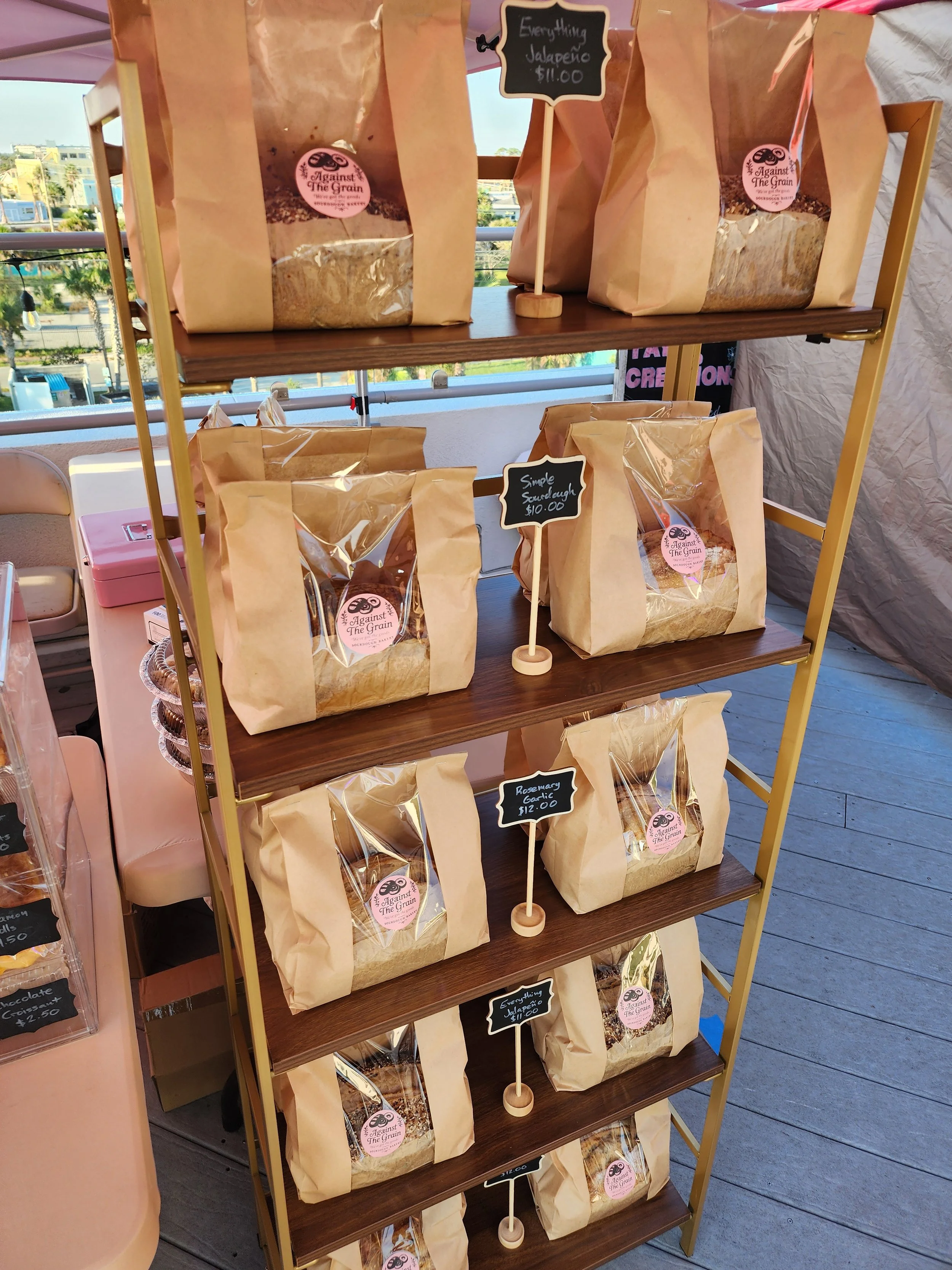 A wooden display shelf with multiple paper bags filled with grain, each labeled with different flavors and prices. There are small signs indicating flavor options like 'Simple Sourough $10.00' and 'Everything Jalapeño $11.00'. The bags have pink and 