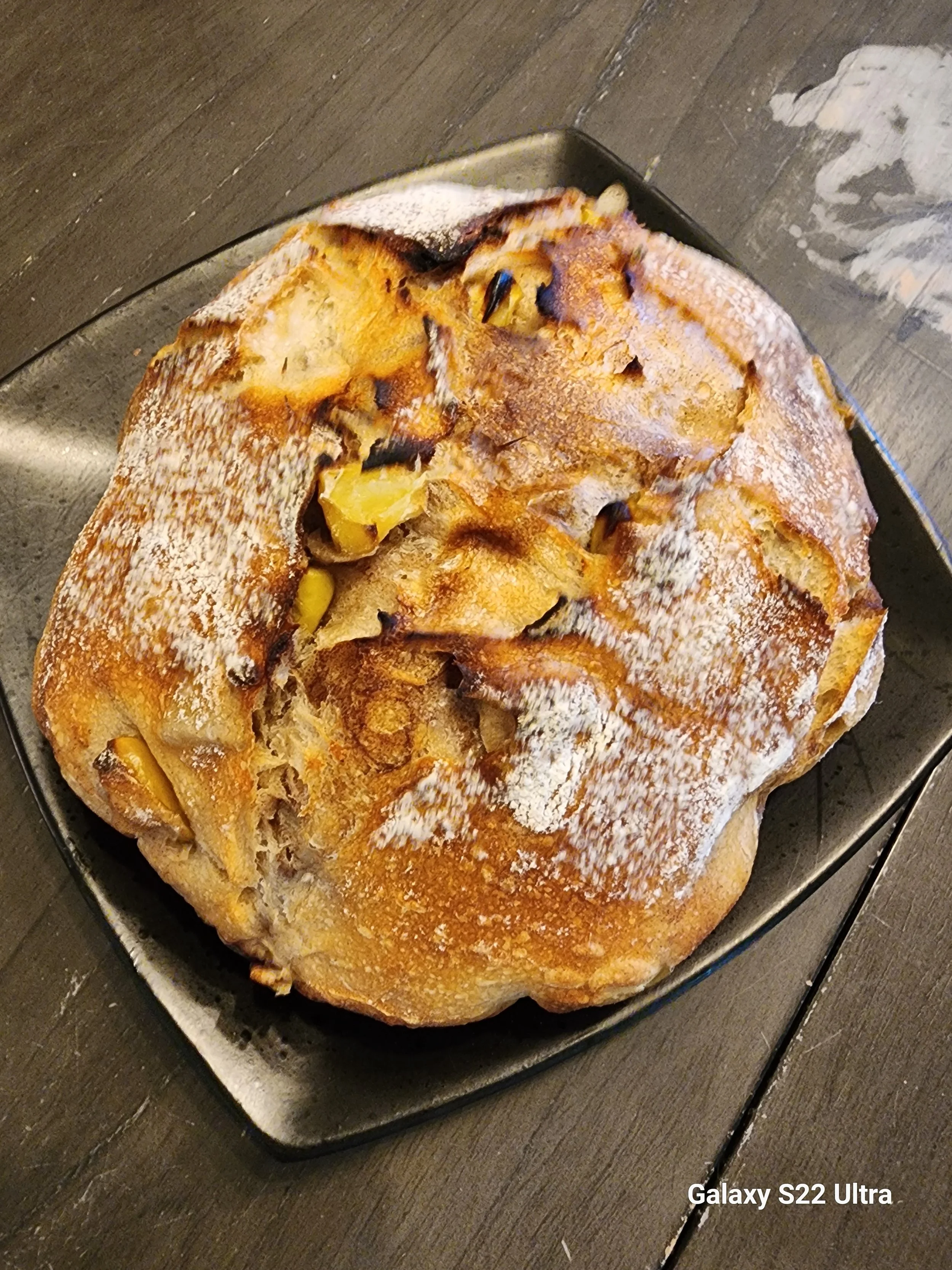 A freshly baked loaf of bread with a golden-brown crust, dusted with flour, placed on a metal tray on a dark wooden surface.