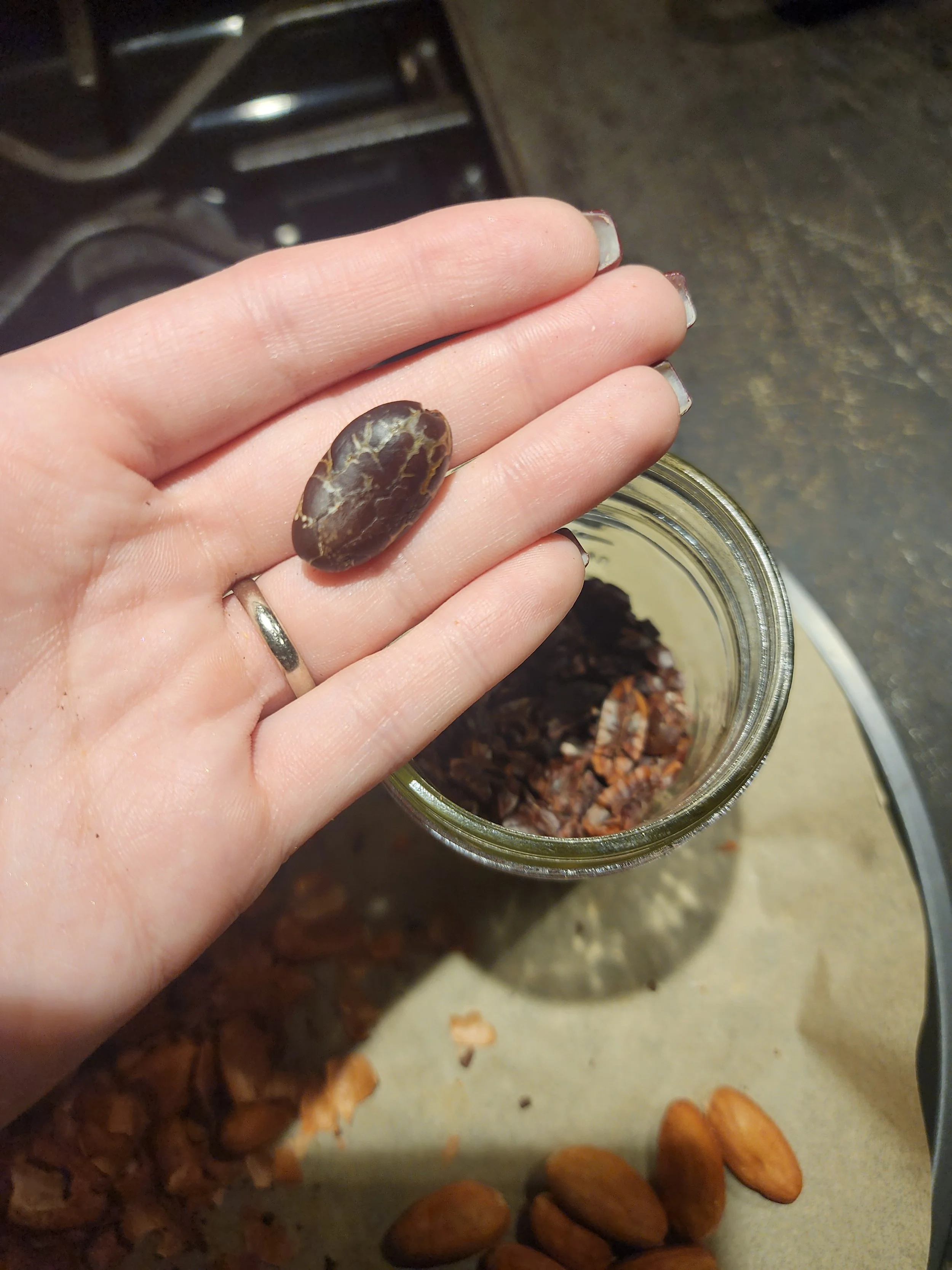A person's hand holding a dark, oval-shaped, textured stone over a jar filled with small rock fragments. The background shows a work surface with scattered rocks and almonds on the lower edge.