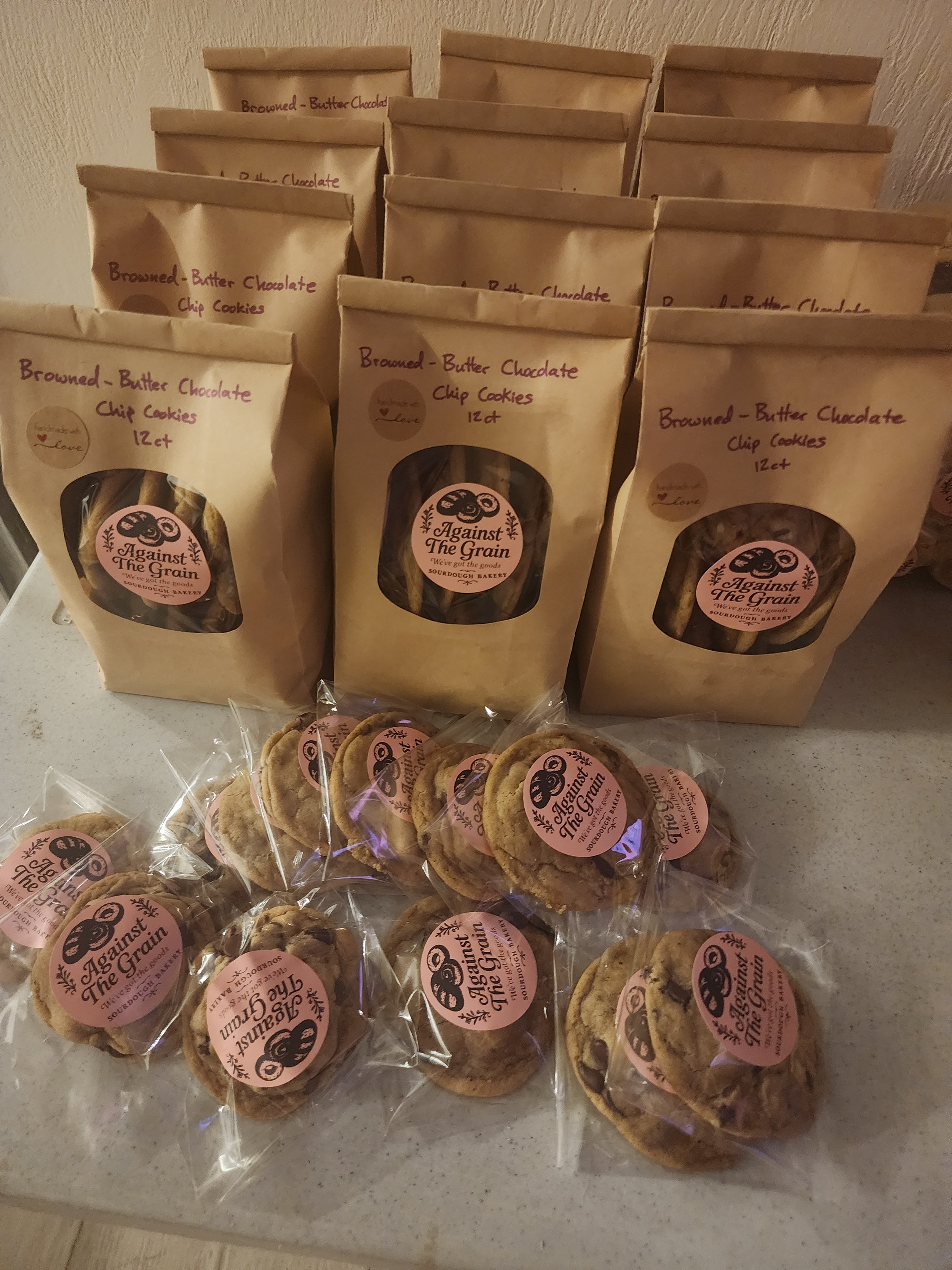 Bags of homemade butter chocolate chip cookies labeled 'Against The Grain' on a countertop, some cookies in front of the bags in plastic wrapping.
