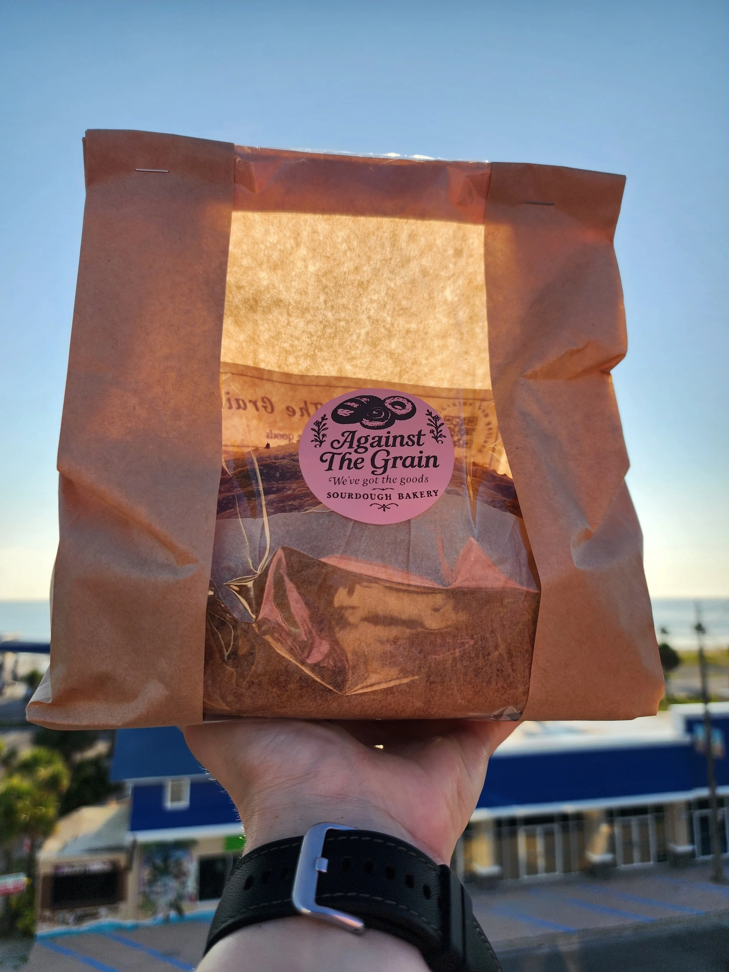 Person holding a paper bag with branding from Against The Grain Bakery, set against a clear sky and a building with blue accents in the background.
