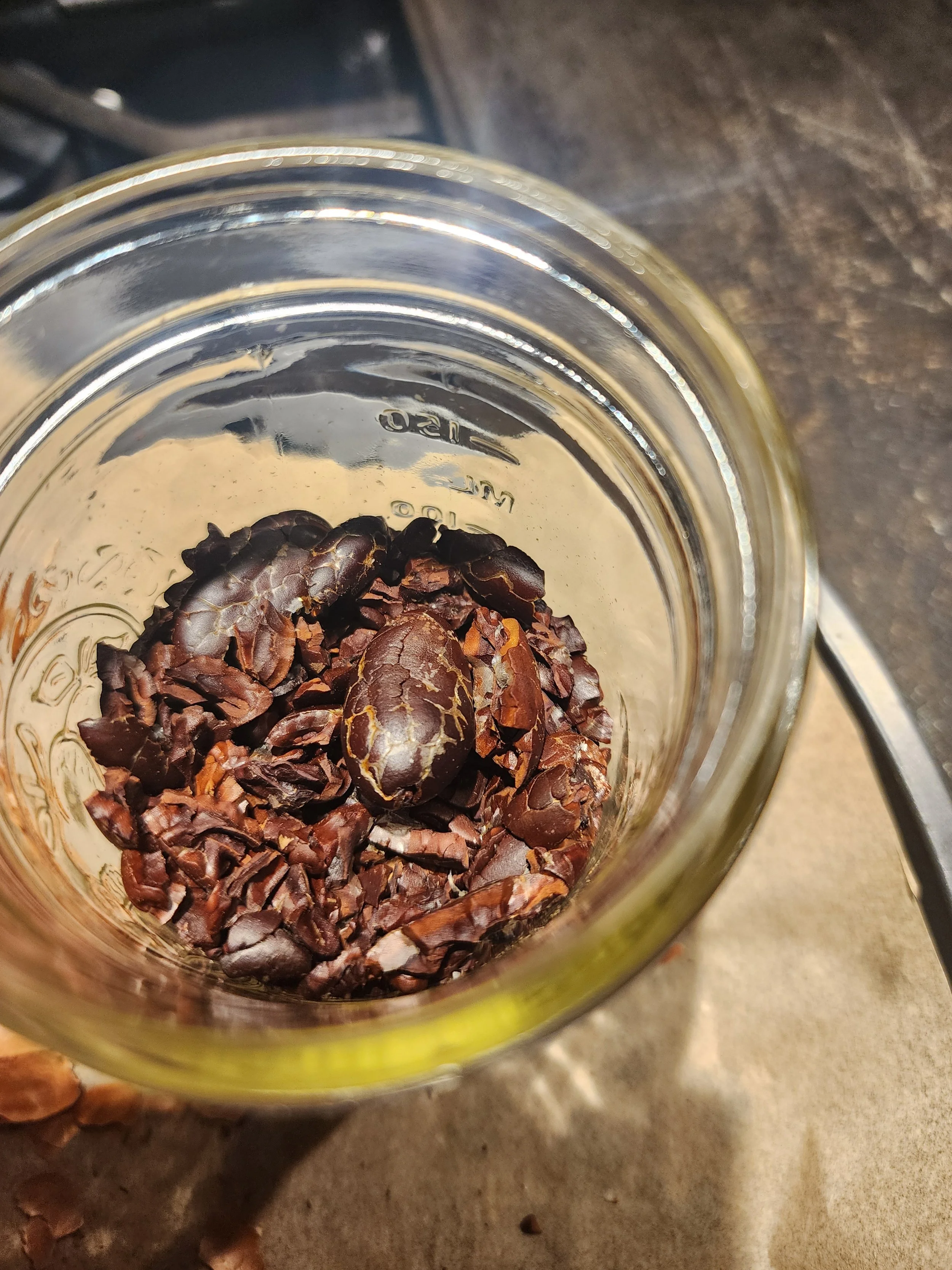 Close-up of a glass jar with small, dark brown, cracked cacao nibs inside, placed on a counter or table surface.