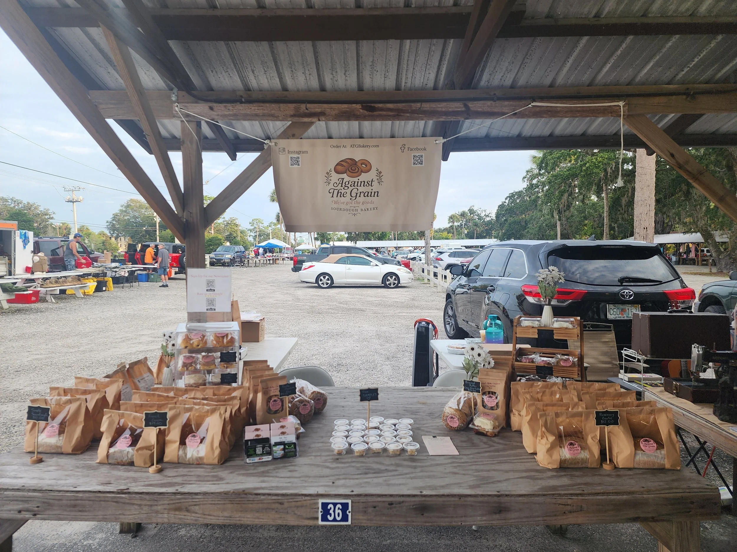 Market stall selling baked goods such as bread, cookies, and muffins at an outdoor event with cars and people in the background.