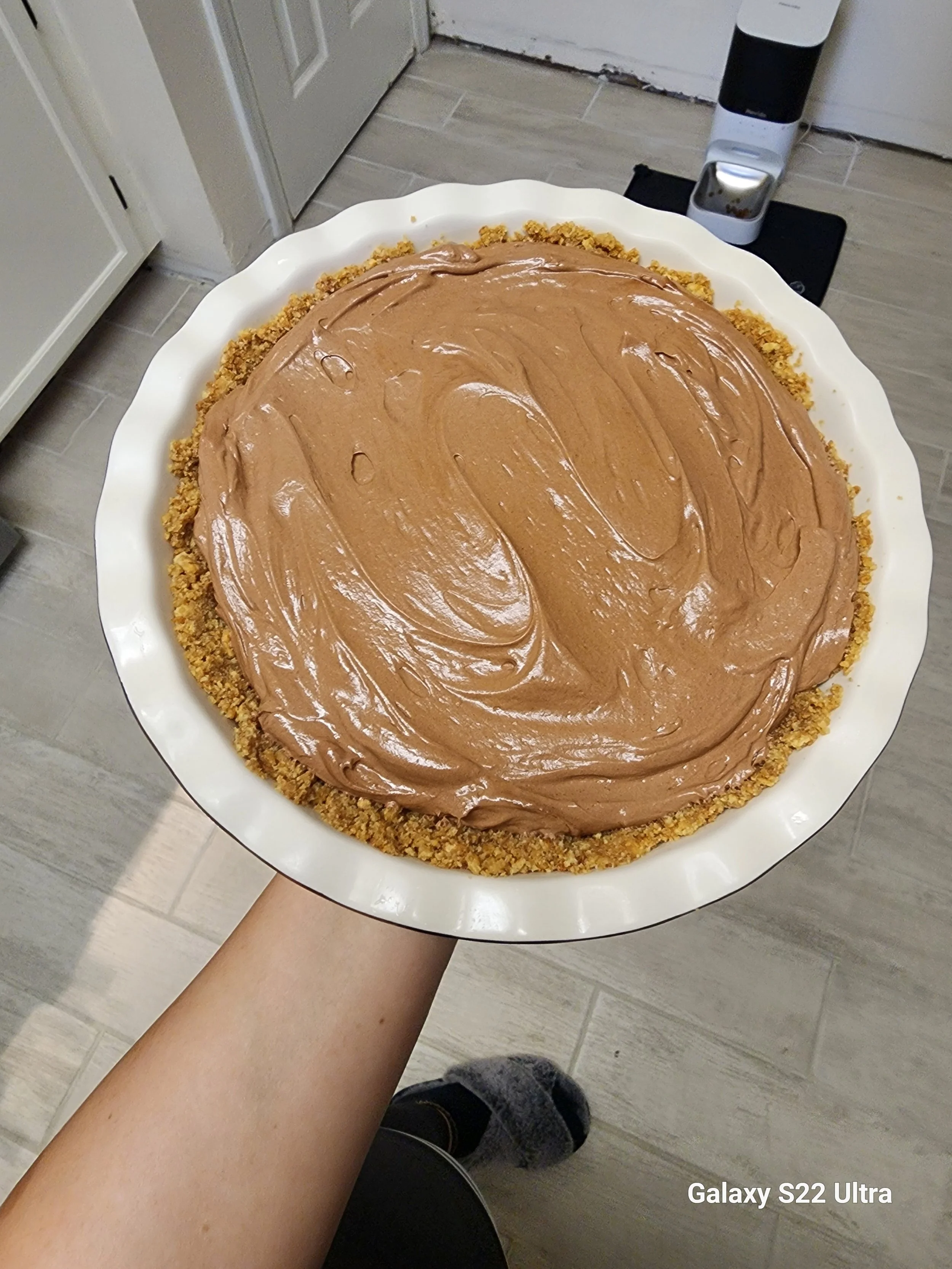 A person holding a large white plate with a graham cracker crust, filled with a smooth chocolate mousse or pie filling, on a kitchen floor with a pet food dispenser in the background.