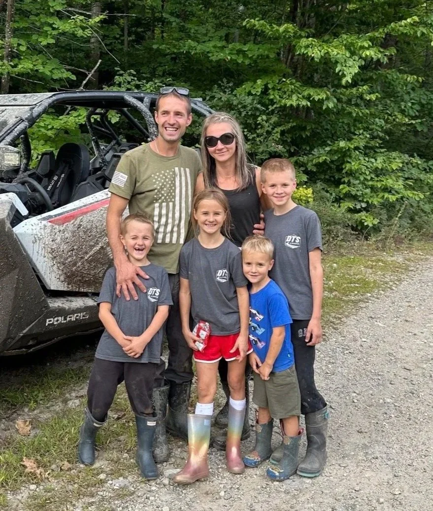A group of six people, including two adults and four children, standing outdoors near a muddy off-road vehicle in a forested area, all smiling at the camera.