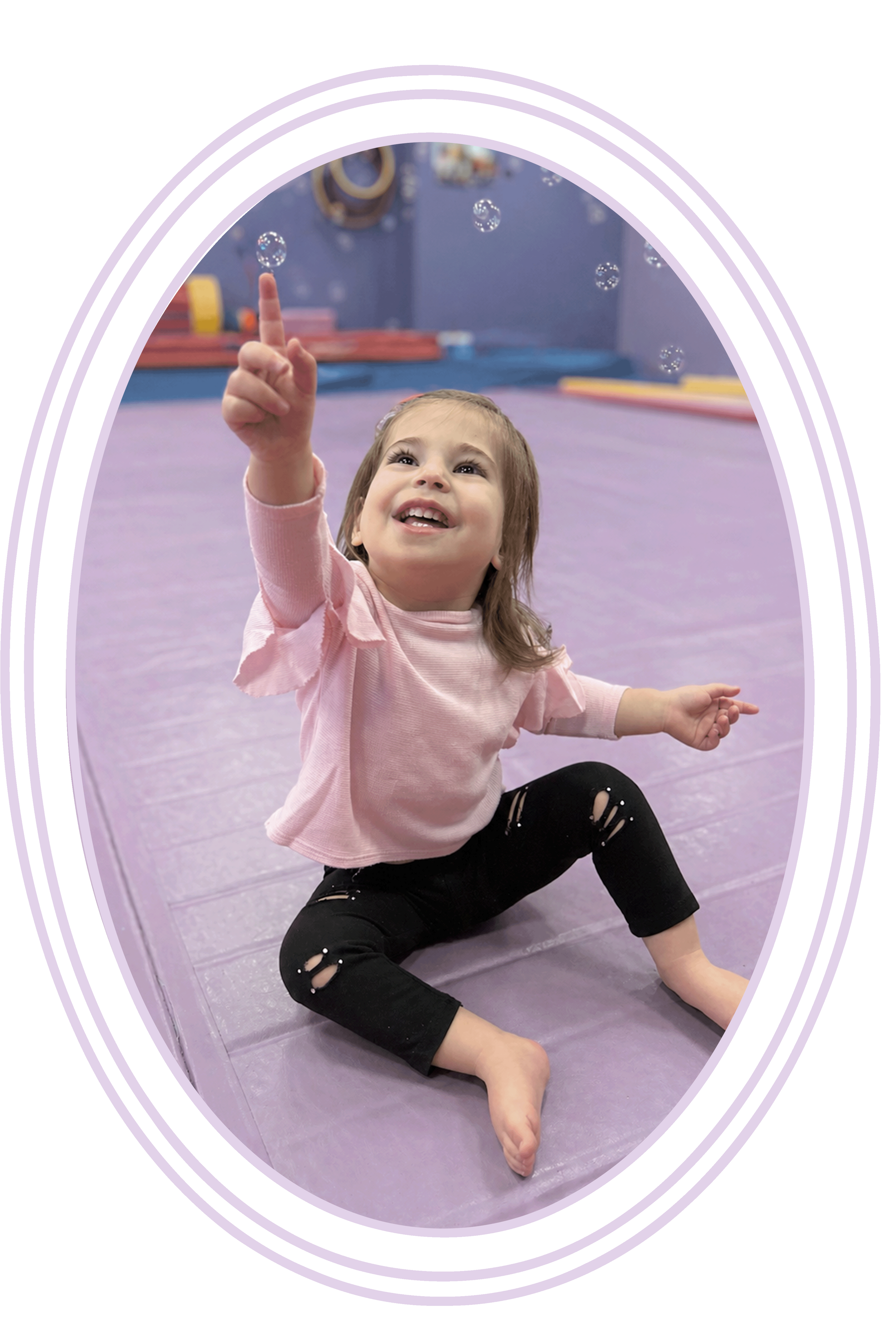 Young girl with brown hair smiling and pointing upward while sitting on a purple floor in a dance or gym studio with pastel-colored walls and equipment in the background.