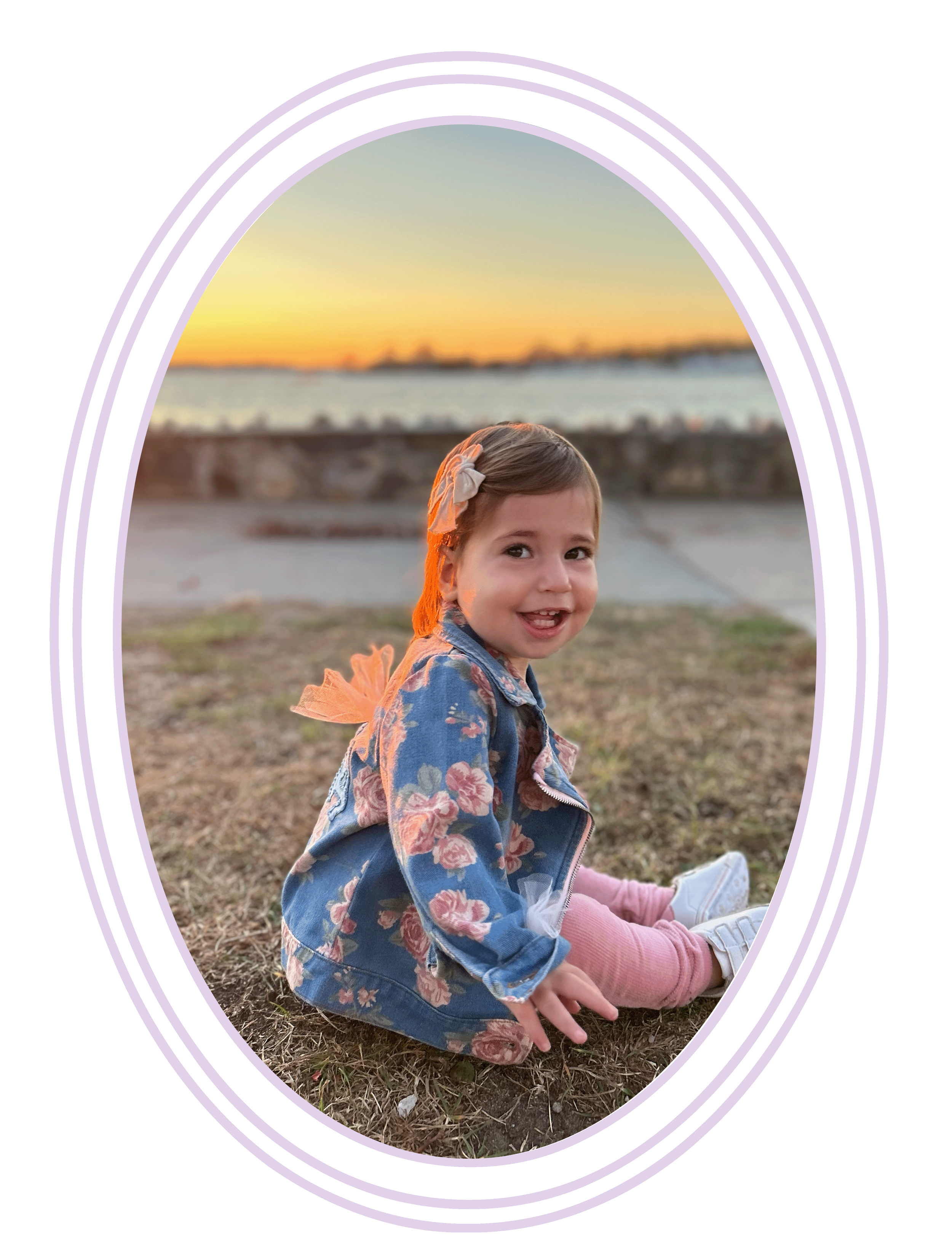 A smiling young girl sitting on the ground outdoors during sunset, wearing a blue floral jacket, pink shoes, and white sneakers, with her hair tied with a beige bow.