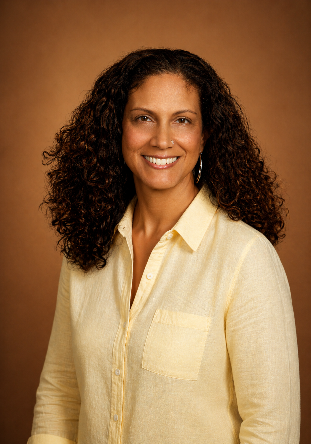 A woman with curly dark hair, wearing a light yellow button-up shirt, smiling against a brown background.