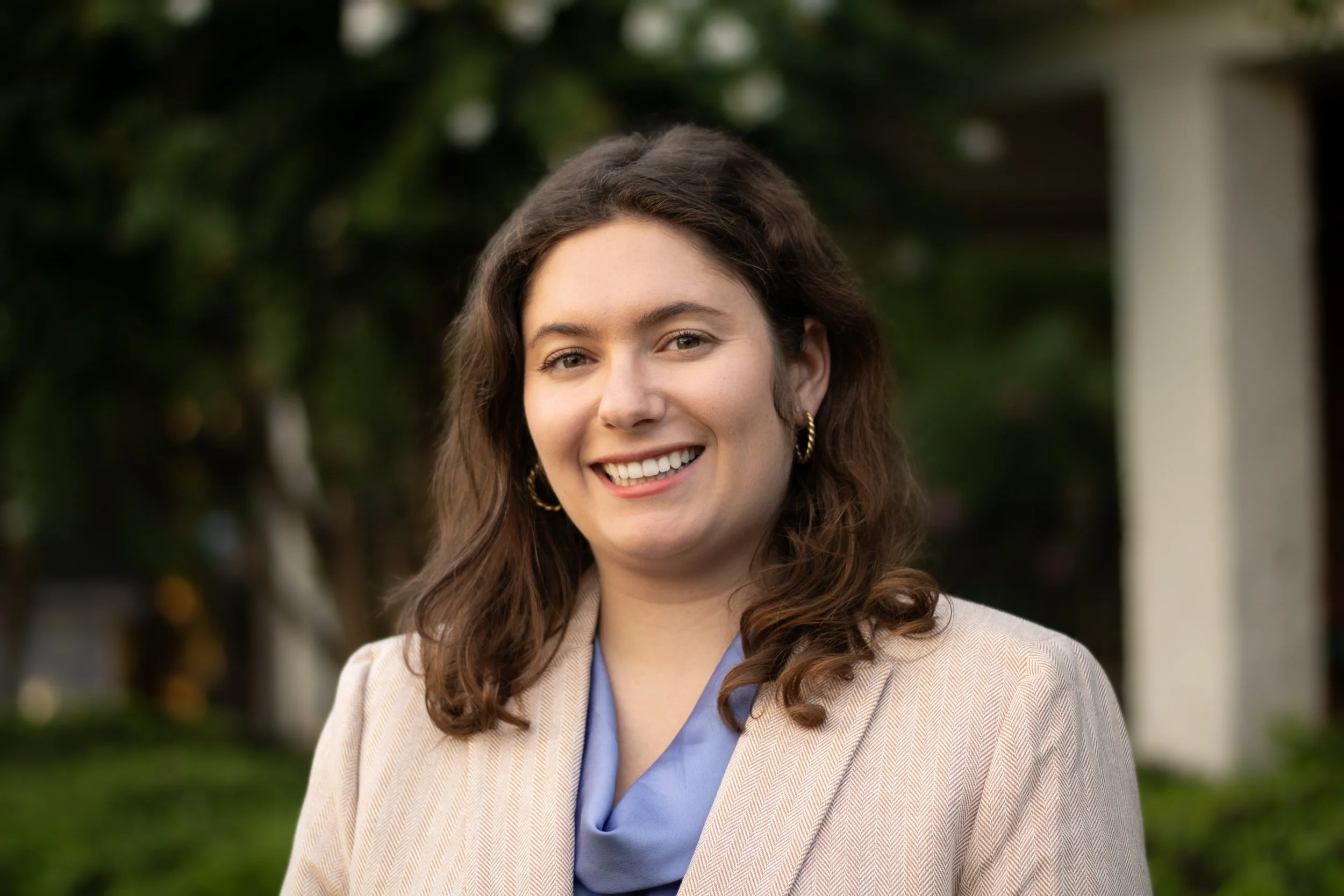 Portrait of a smiling woman with brown hair wearing a beige blazer and gold hoop earrings outdoors.