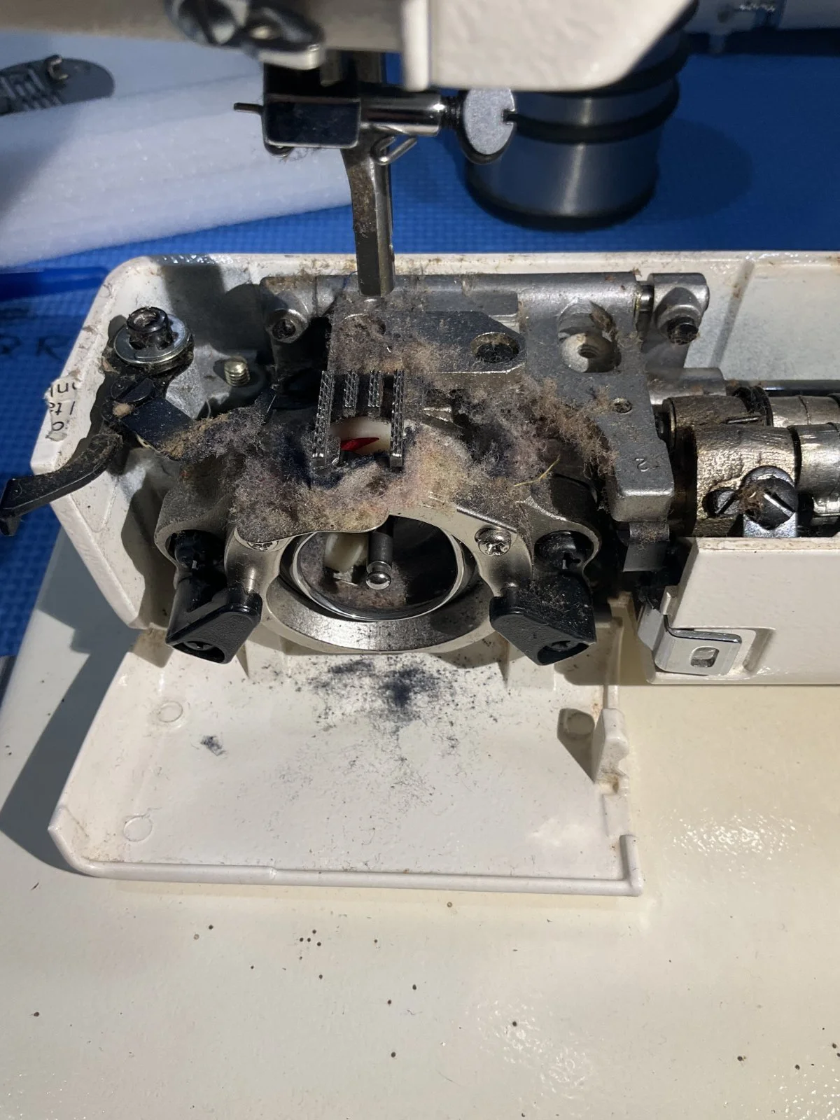 A close-up of an internal mechanical part of a sewing machine with lint, dust, and debris, showing gears, screws, and metal components.