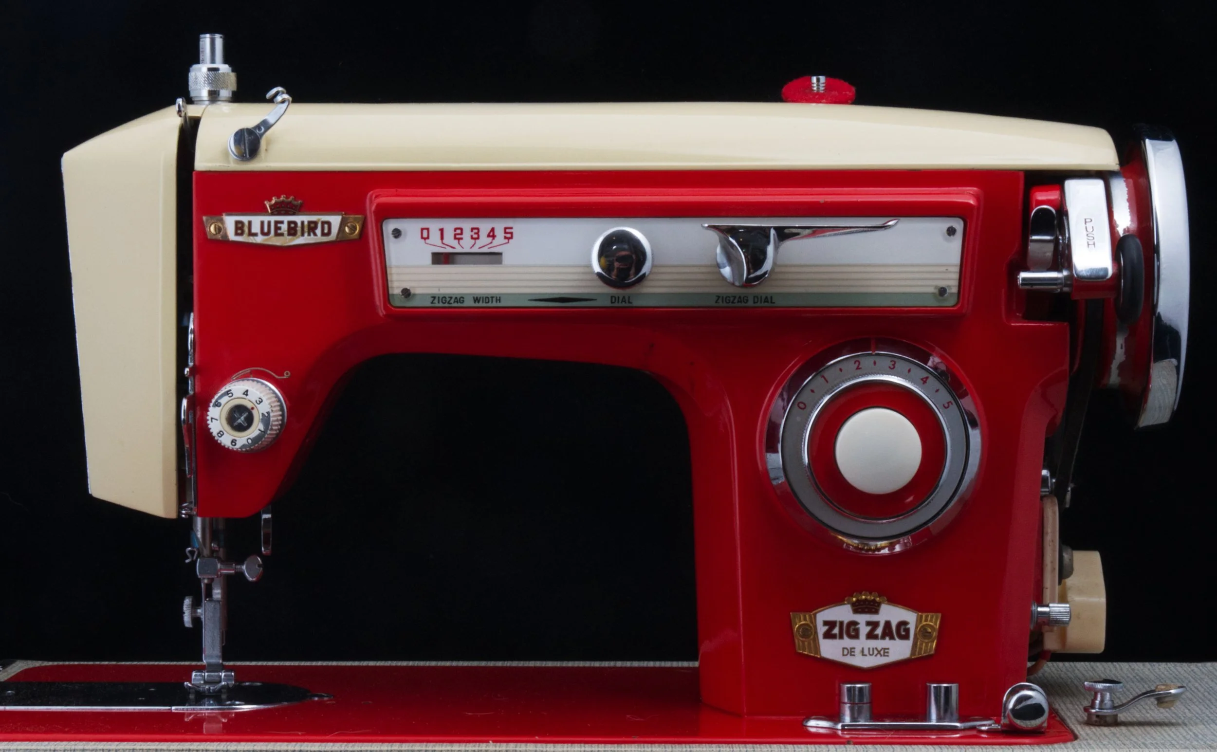 A vintage Bluebird Zig Zag sewing machine with red and cream-colored body and metal components, set against a black background.