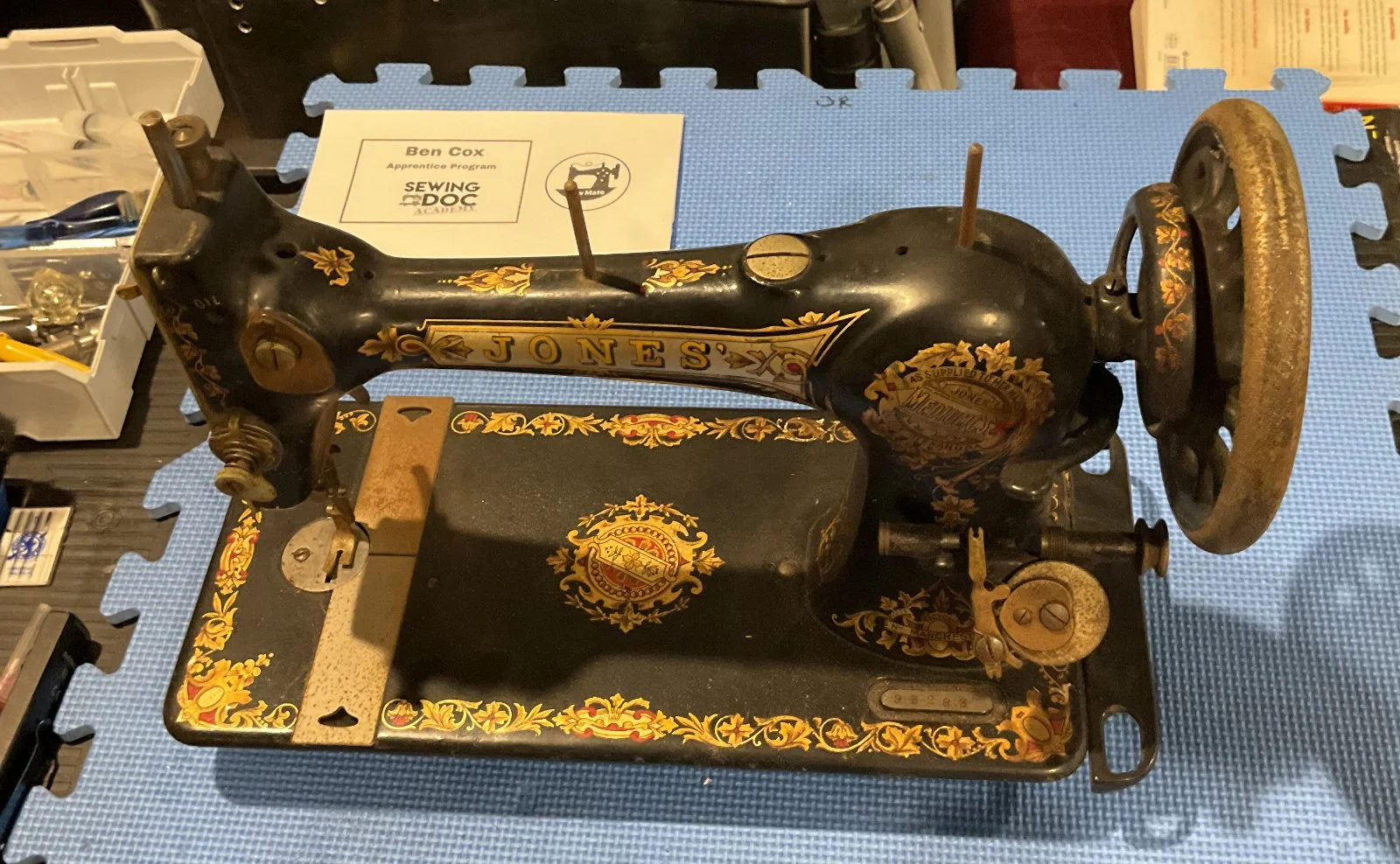 Vintage black and gold Jones sewing machine with ornate decorative patterns, placed on a blue foam mat, with a white box and document behind it.