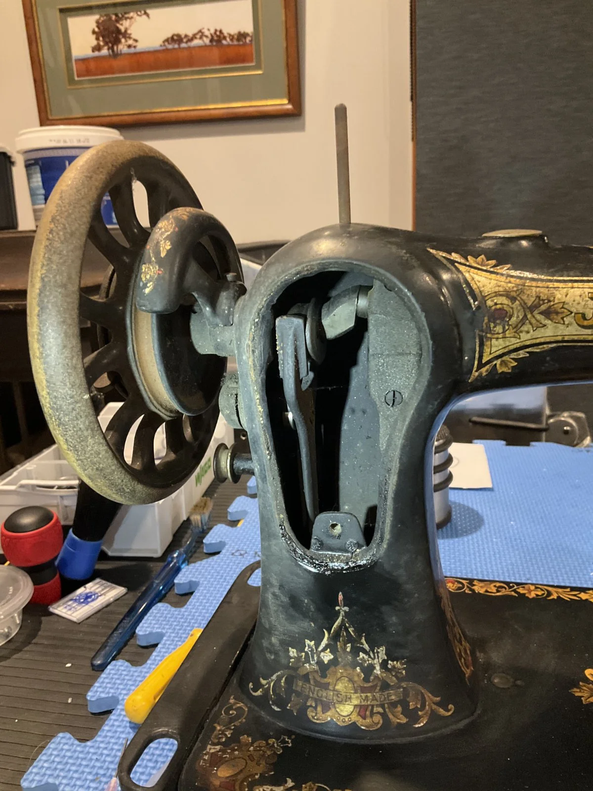 Close-up of an antique Jones sewing machine before restoration, with exposed internal mechanisms, and a large wheel with rust, situated on a worktable with tools and supplies, in a room with framed picture and other furniture.