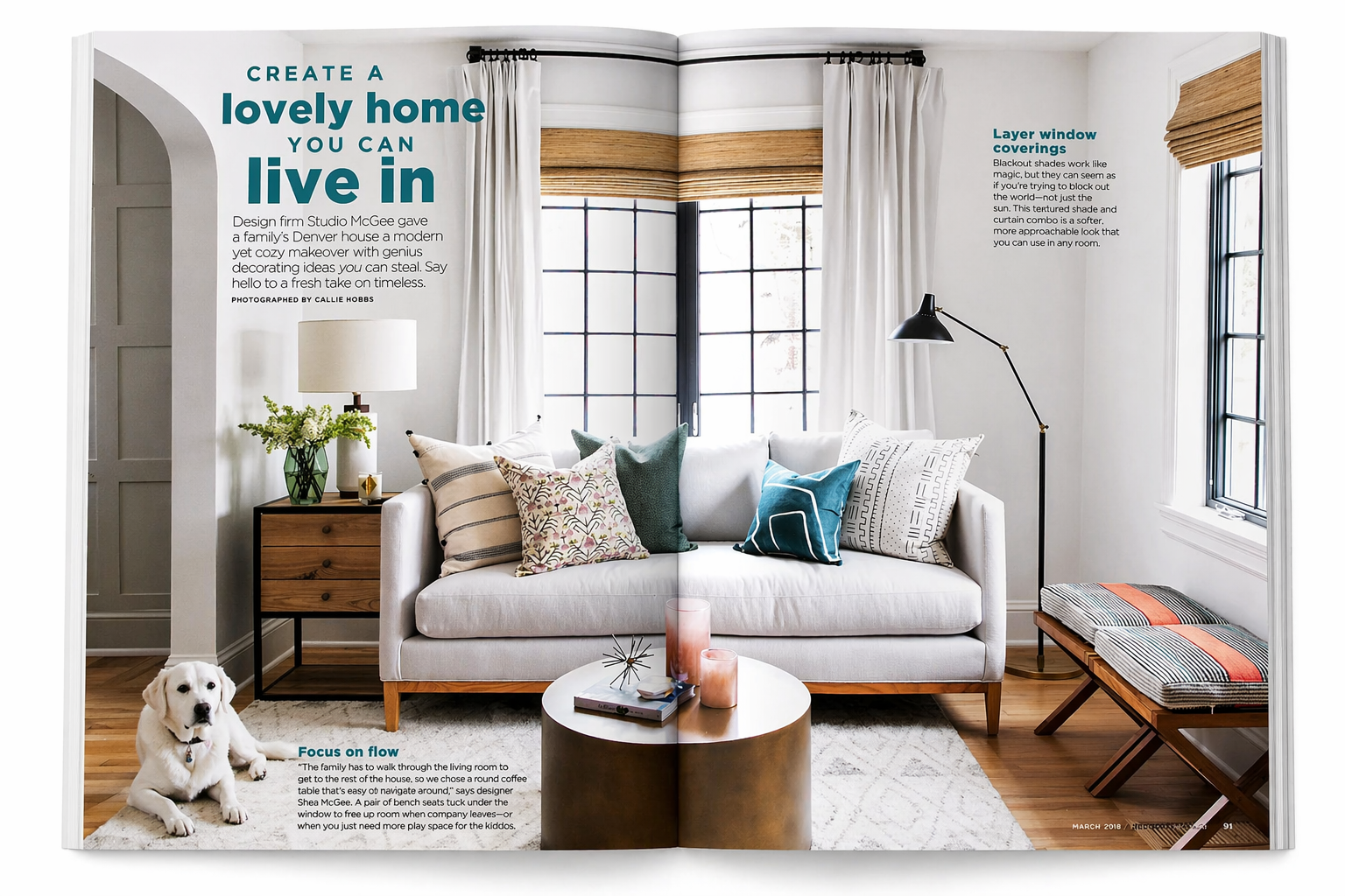 Bright living room with a white sofa adorned with several colorful patterned pillows, a round coffee table with candles and books, a wooden bench with a striped cushion, a side table with a lamp and green plant, a black floor lamp, and large windows 