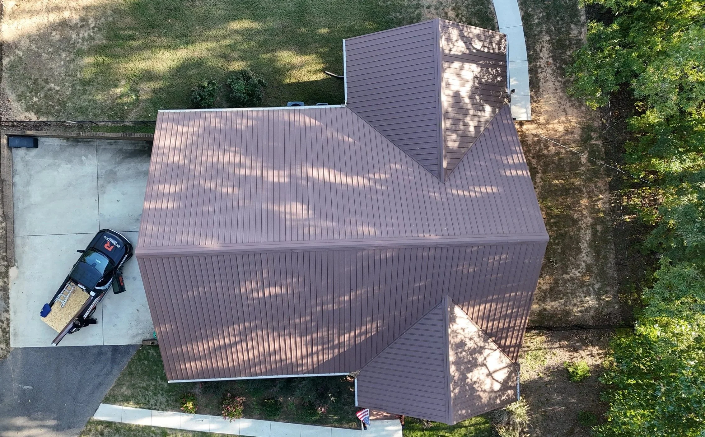 An aerial view of a residential backyard showing a house with a brown metal roof, a concrete driveway with a black pickup truck with a ladder in the bed, a small shed, green grass, trees, and a sidewalk.