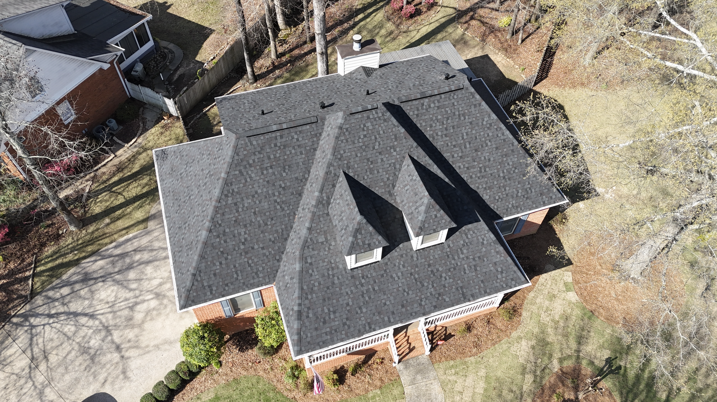 Aerial view of a house with a gray shingled roof, multiple peaks, and a chimney, surrounded by trees with sparse foliage and neighboring houses, casting shadows on the yard.