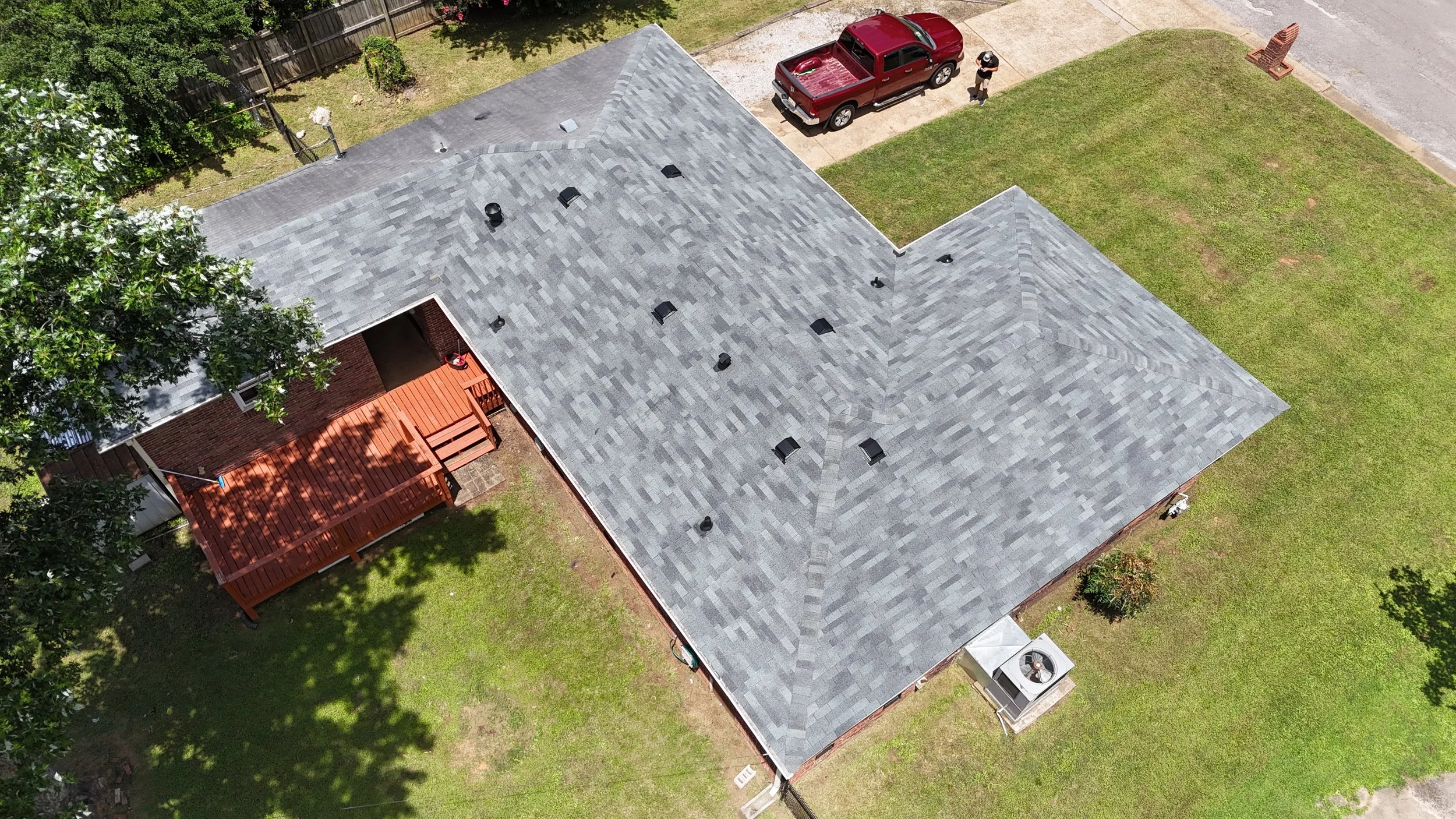 An aerial view of a house with a gray shingle roof, a wooden deck with stairs, a red pickup truck parked on the driveway, and a person walking nearby. The yard has green grass, trees, and a brick firepit.