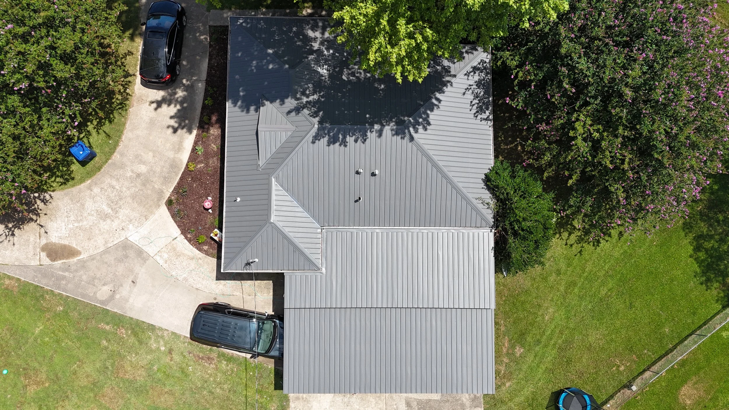 An aerial view of a house with a silver gray metal roof, surrounded by trees, parked cars, and a curved driveway with a blue recycling bin.