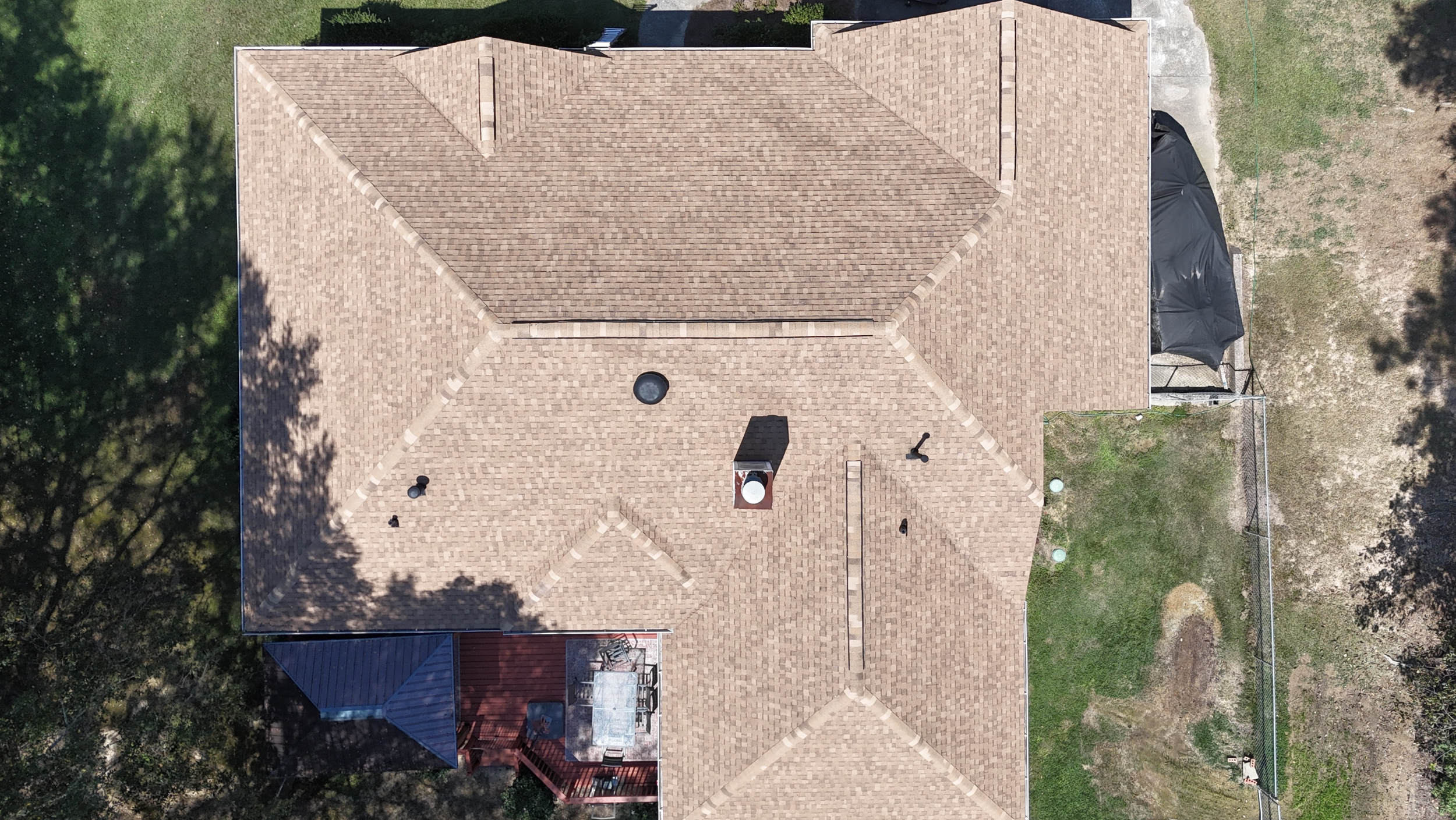 Aerial view of a house with a brown shingle roof surrounded by trees and a fenced yard.