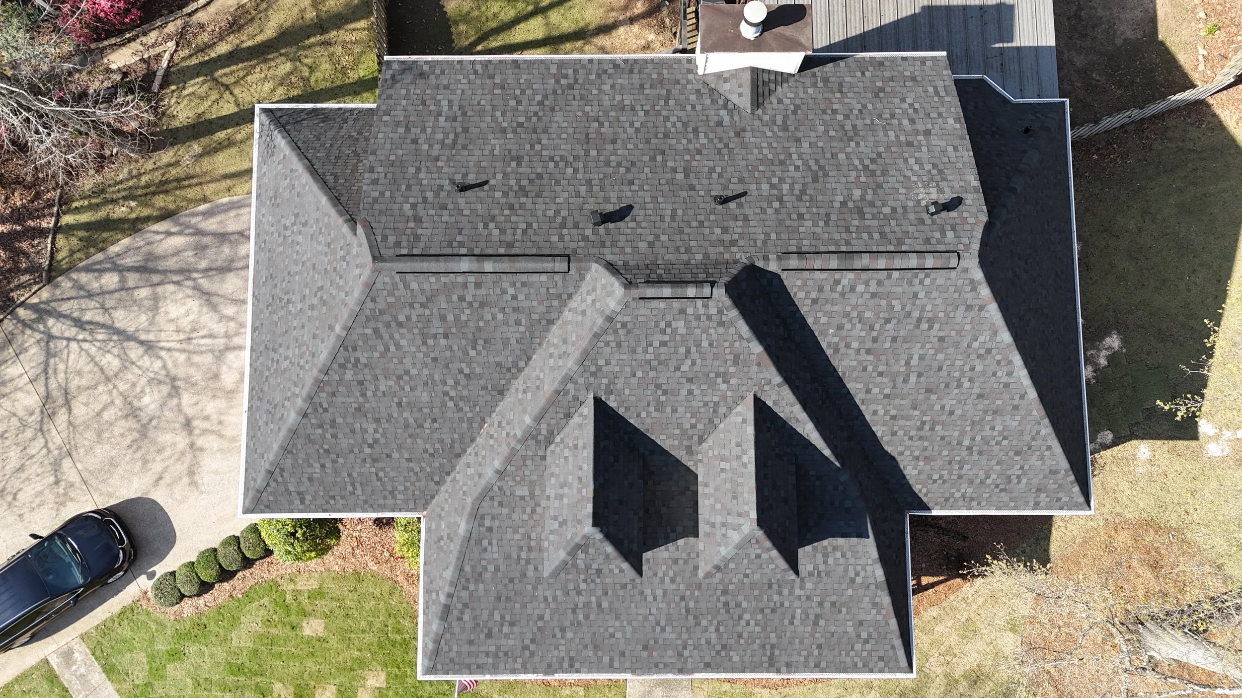 An aerial view of a house with a gray shingle roof, surrounded by trees, grass, and a driveway with parked cars.