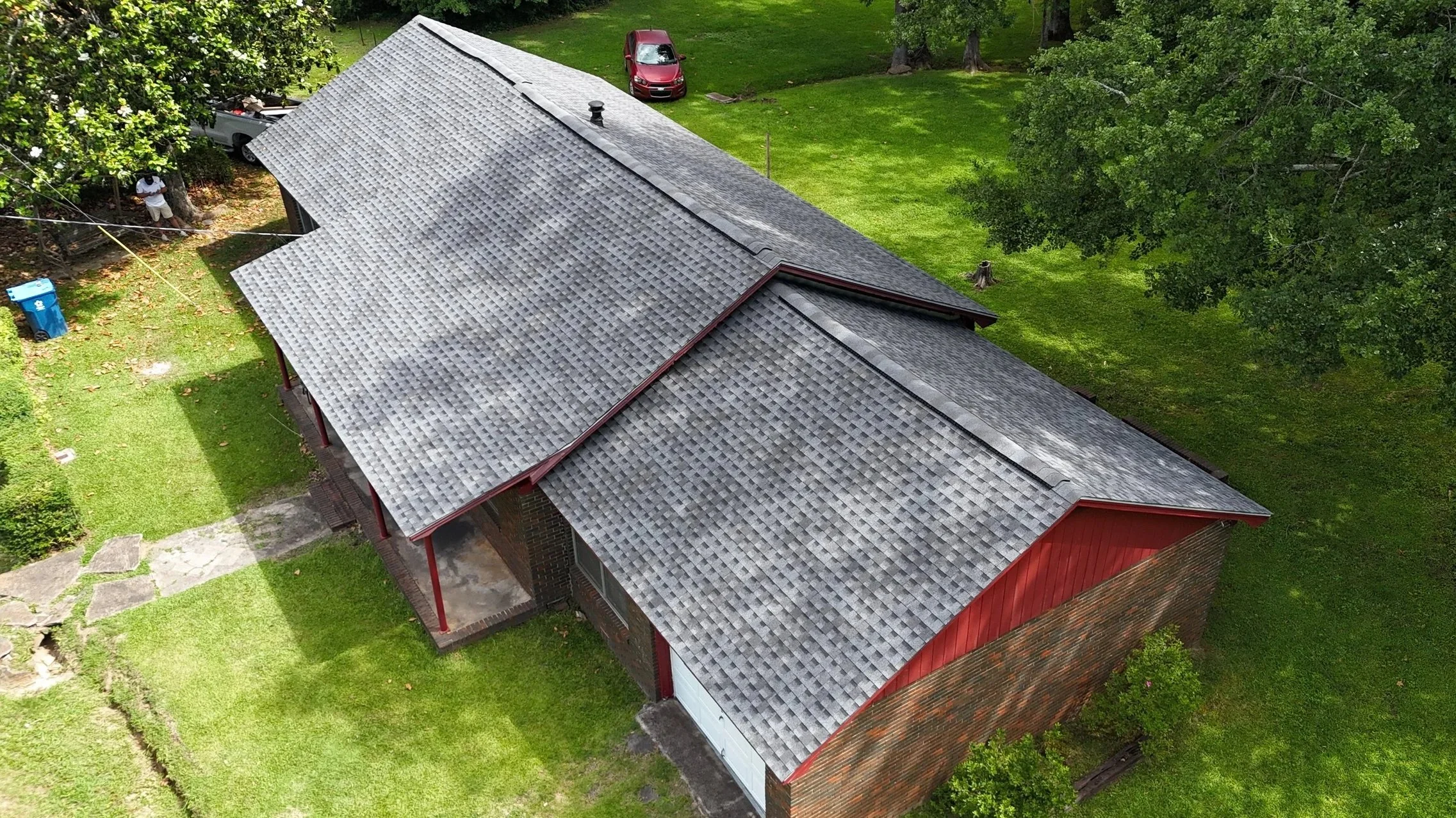 Aerial view of a residential house with a gray shingle roof, surrounded by green trees and grass, with some parked cars in the yard.